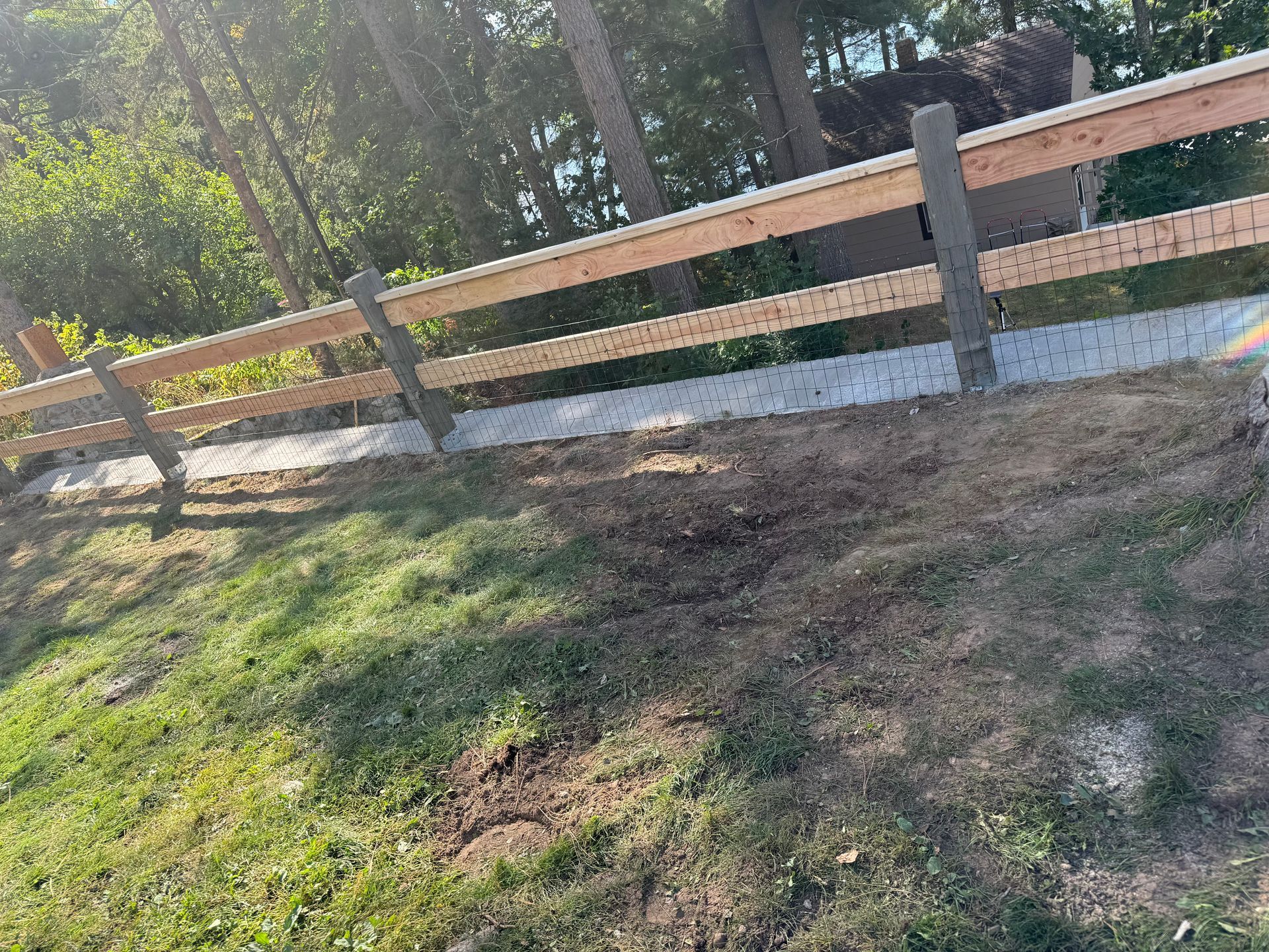 Wooden fence with concrete posts on a grassy slope with trees in the background.