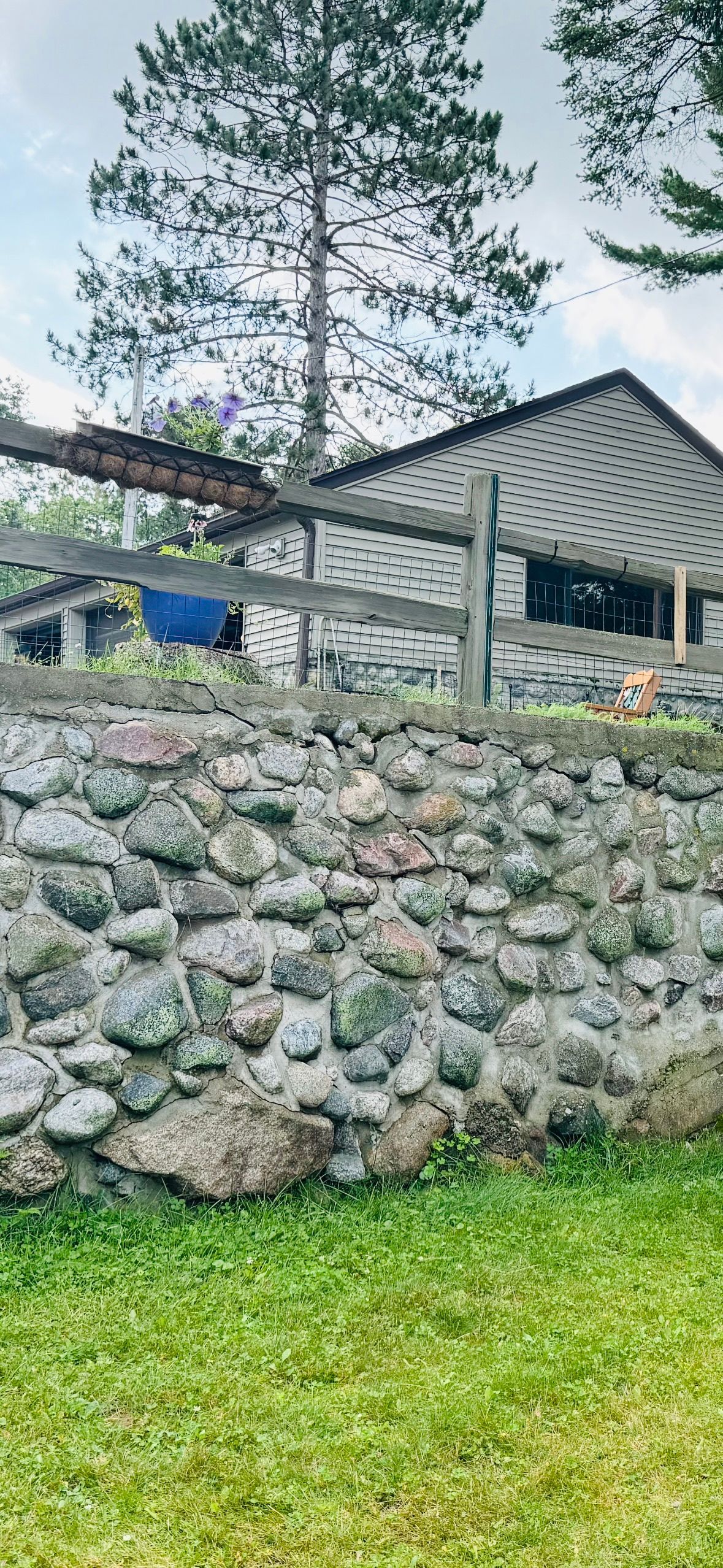 Stone wall with a wooden fence and a house with a patterned facade. Tall trees and green grass.
