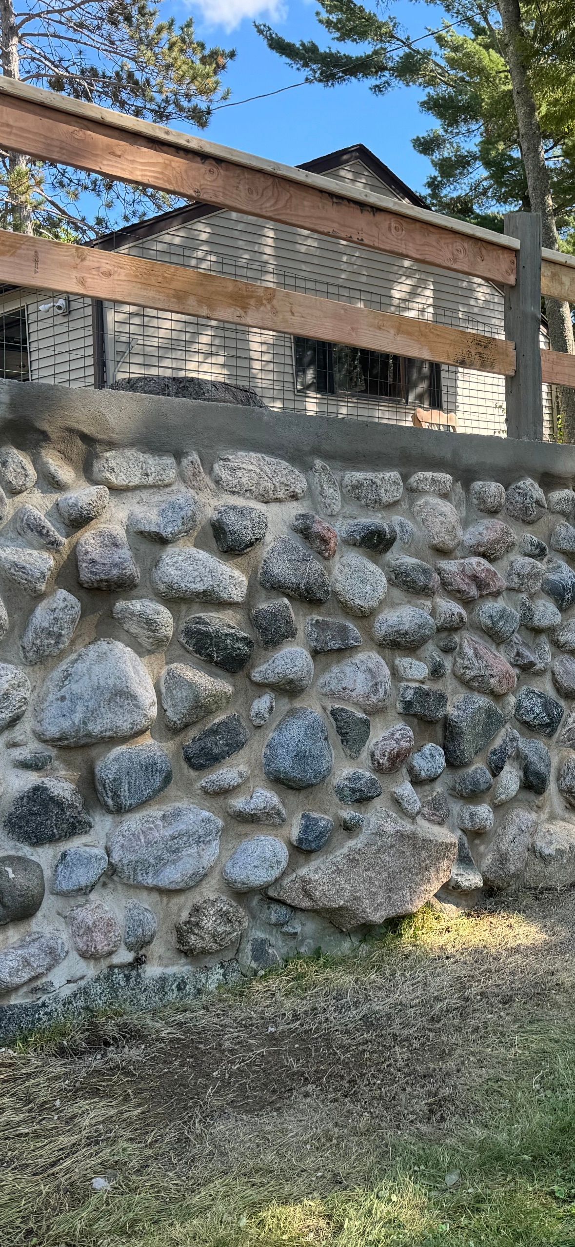 Stone wall supporting a wooden fence, with a building behind it. Green grass at the bottom.