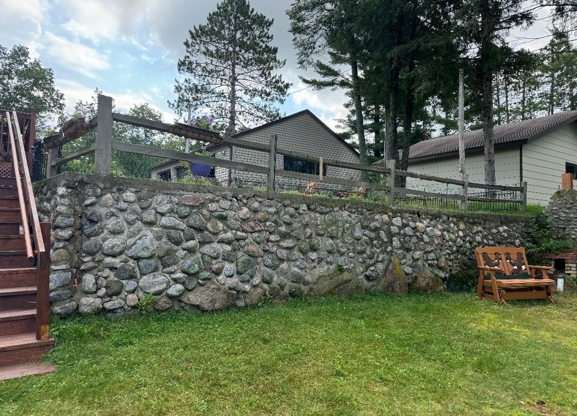 Stone retaining wall supports a wooden fence. A cabin sits beyond the wall, in a grassy area with trees.