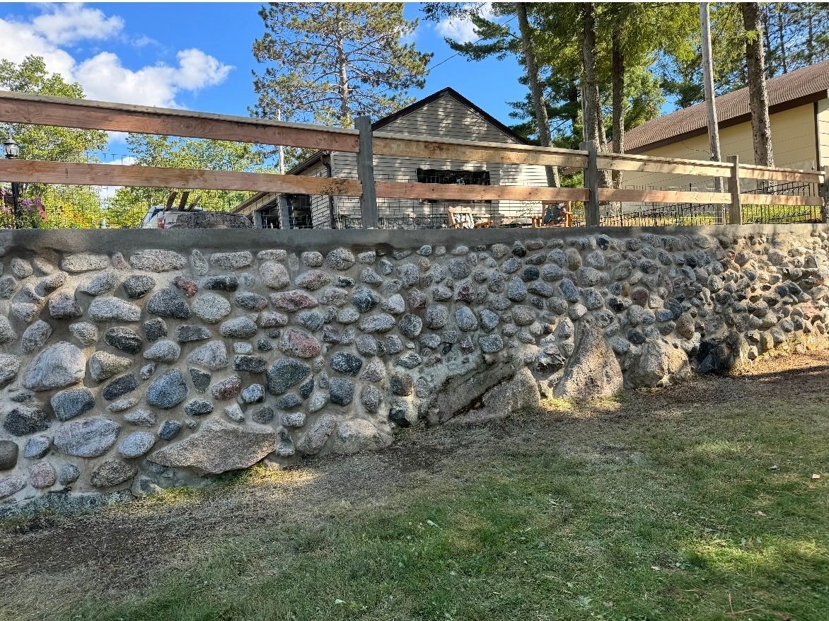 Stone retaining wall with wooden fence and building in the background. Green grass in the foreground, blue sky.
