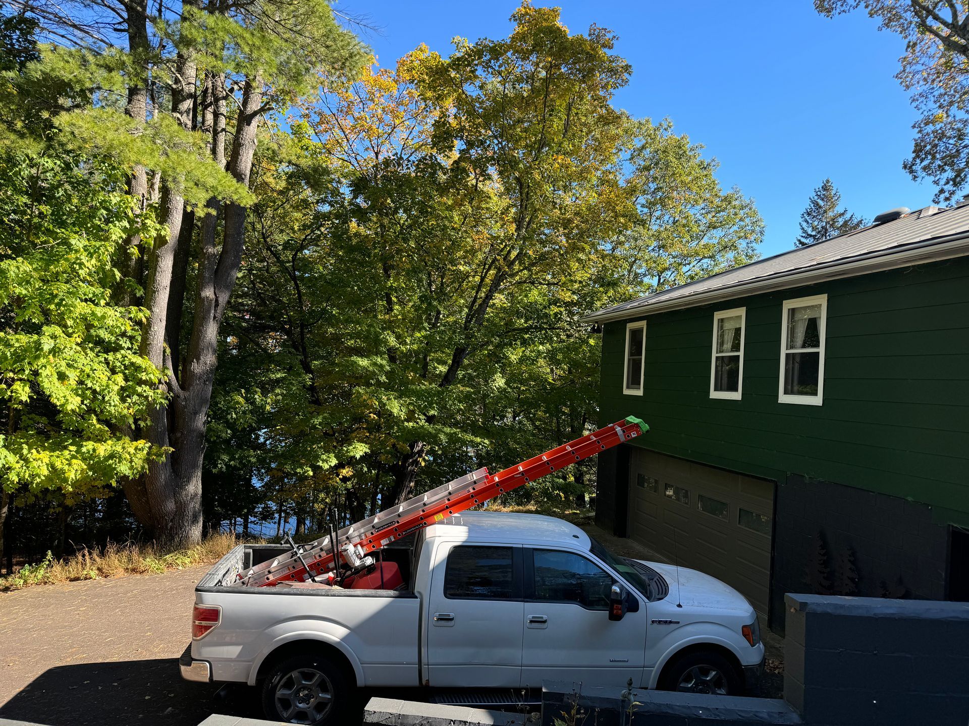 White truck with ladder extending to green house, parked by trees under a blue sky.
