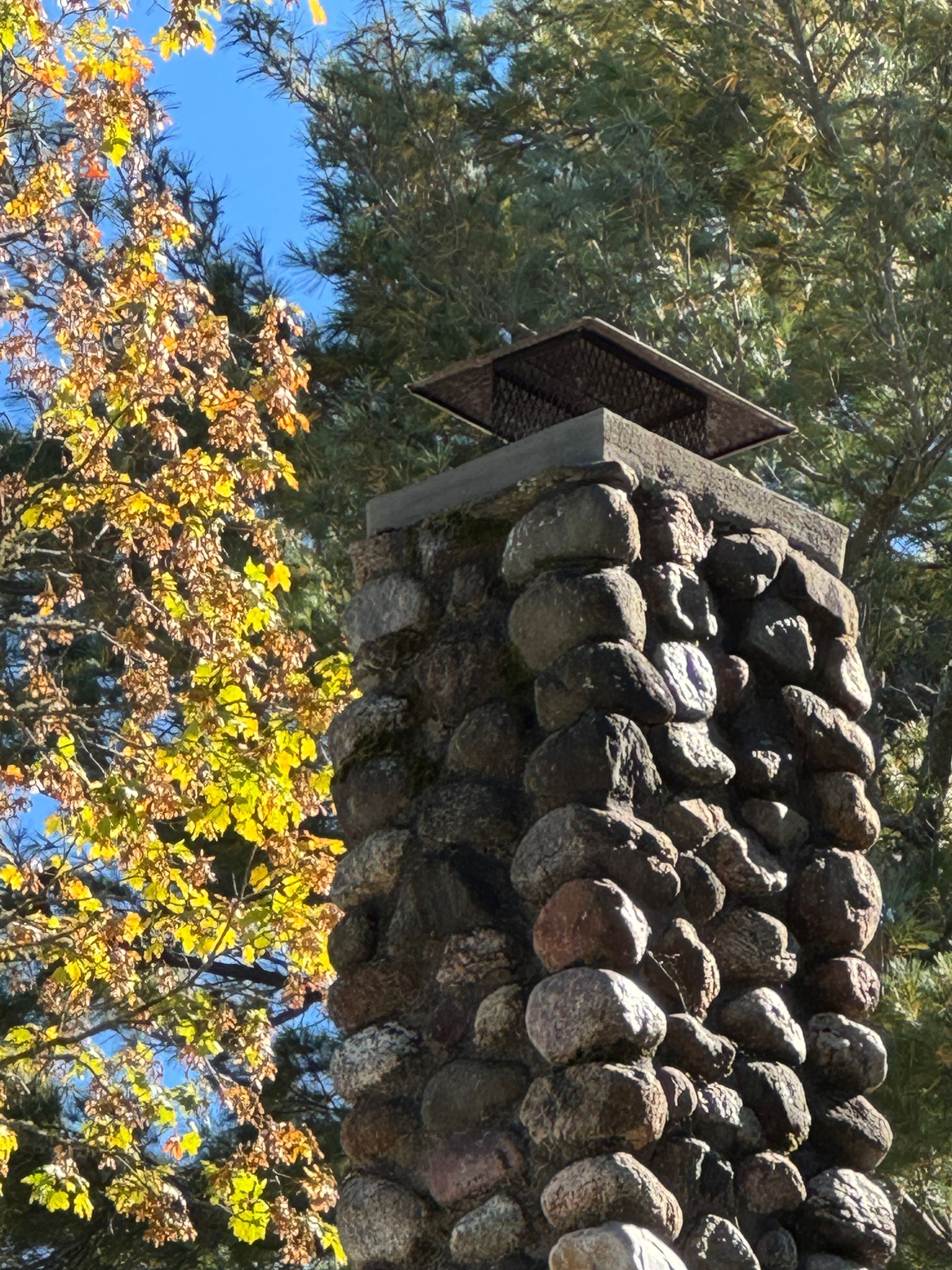 Stone chimney topped with a metal cap, beside autumn tree with yellow leaves.