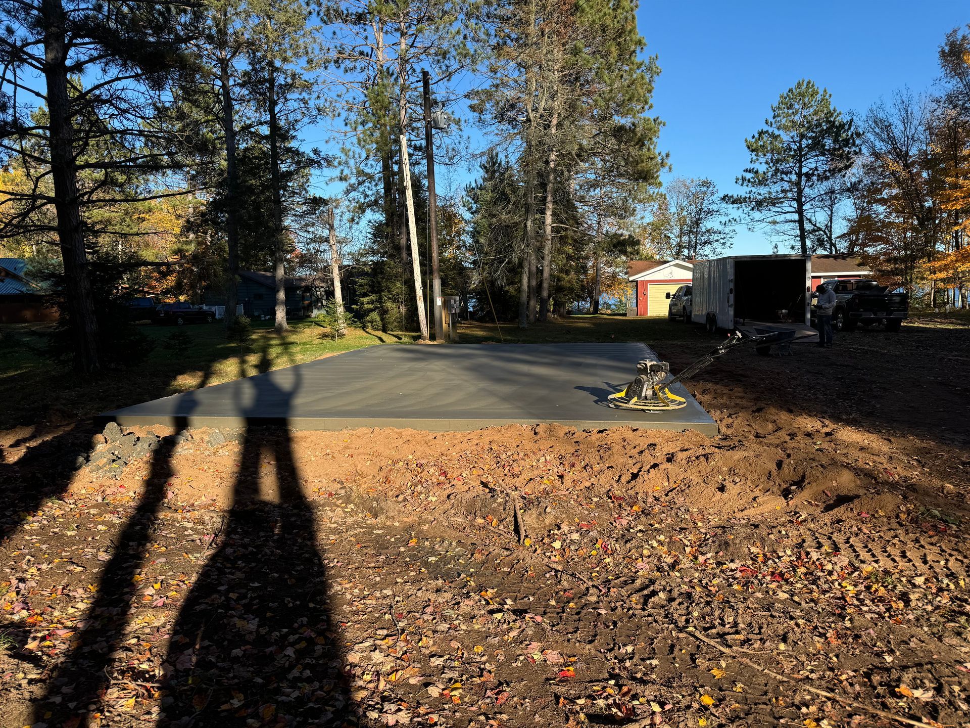 A freshly poured concrete pad sits on a dirt lot, surrounded by trees. A shadow of a person is cast on the ground.