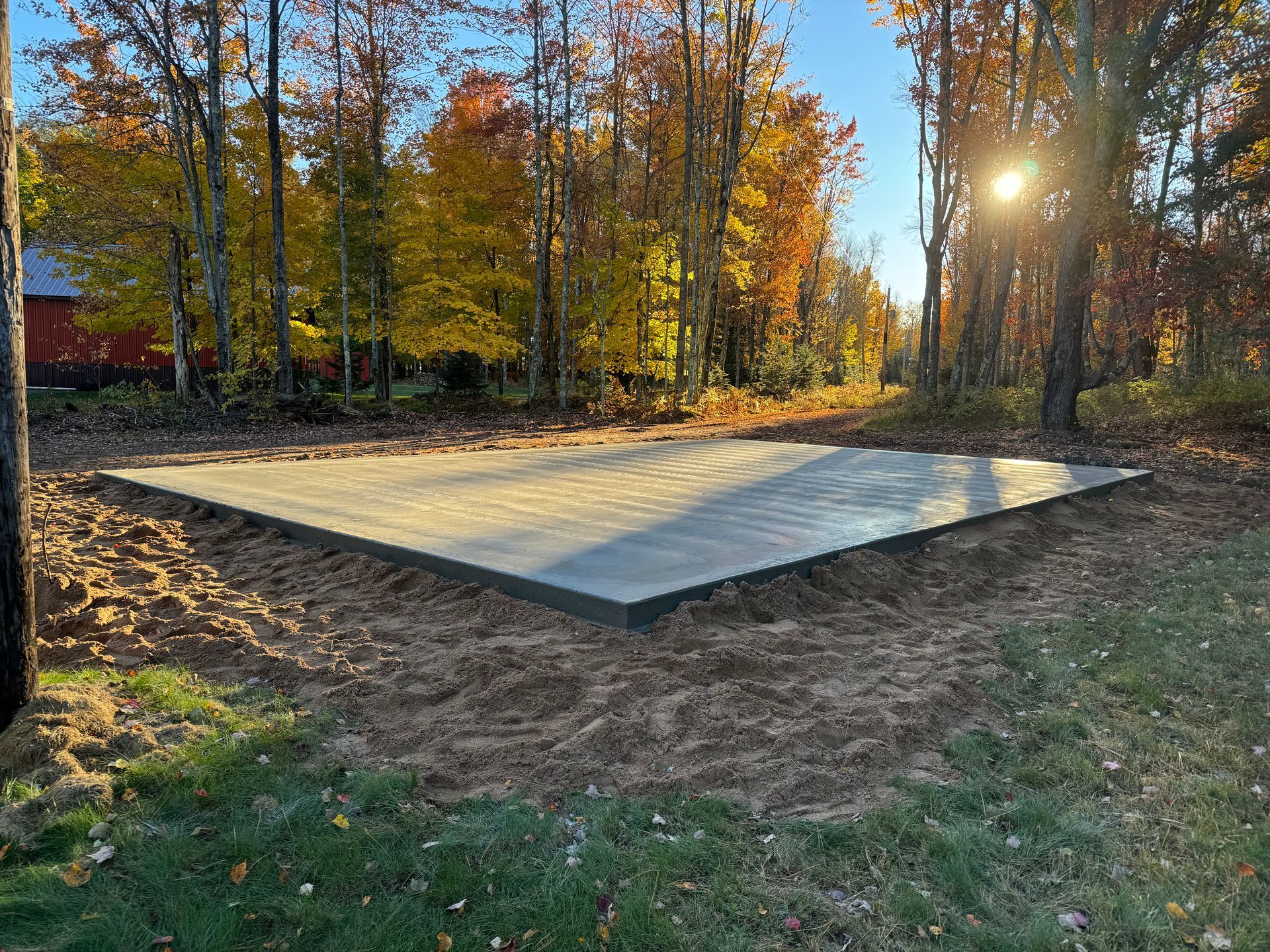 Newly poured concrete slab in a grassy area, surrounded by dirt and autumn trees.