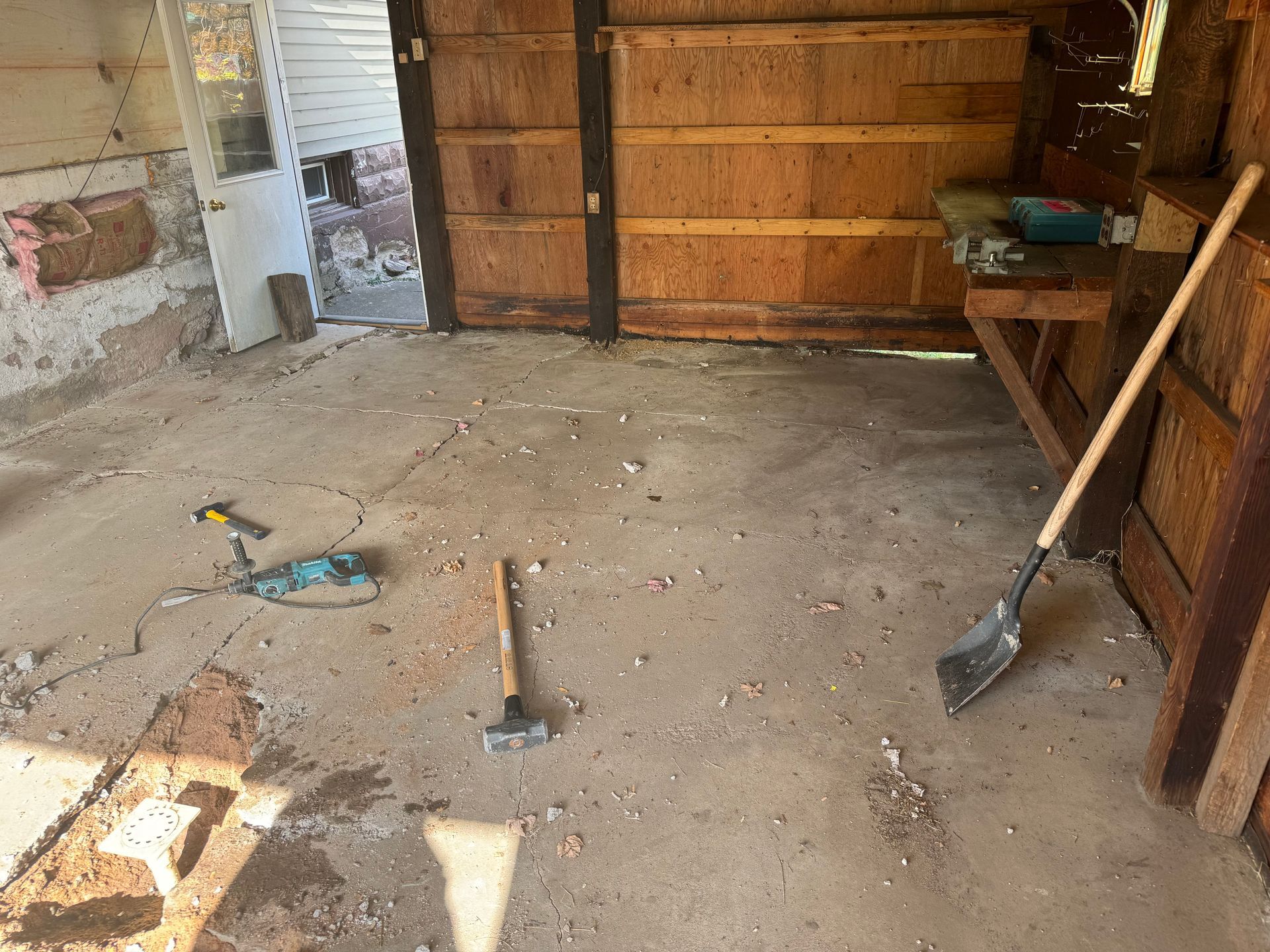 Interior of a shed under construction; concrete floor, wooden walls. Tools, including a shovel and hammer, scattered about.