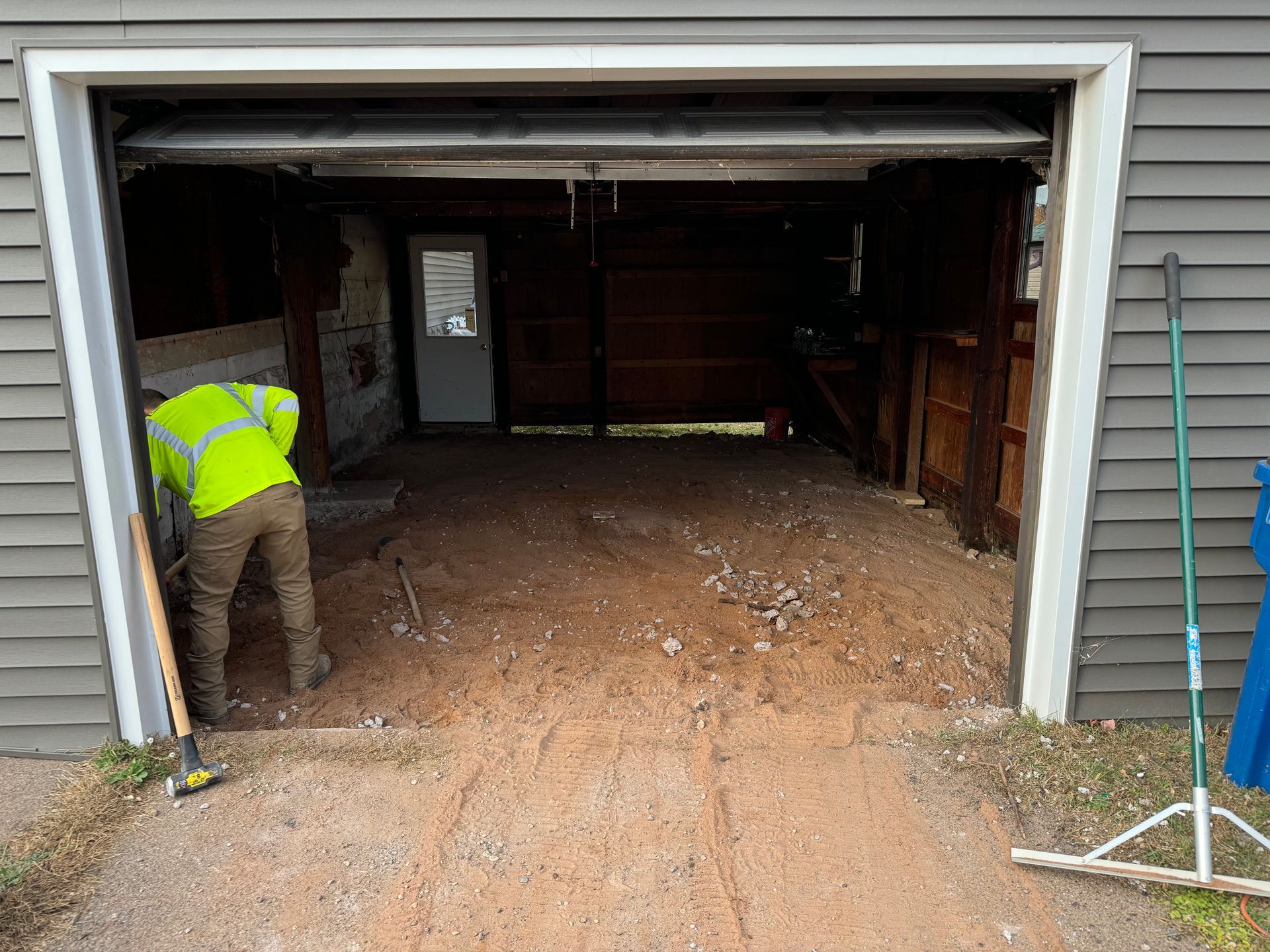 Person in neon vest raking debris from a garage interior. Open garage door, gravel floor, wooden walls.
