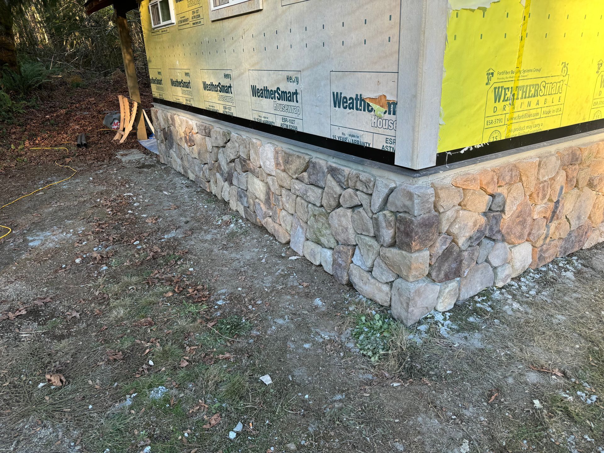 Stone veneer on a building's foundation corner. Green and yellow sheathing above. Natural ground in foreground.