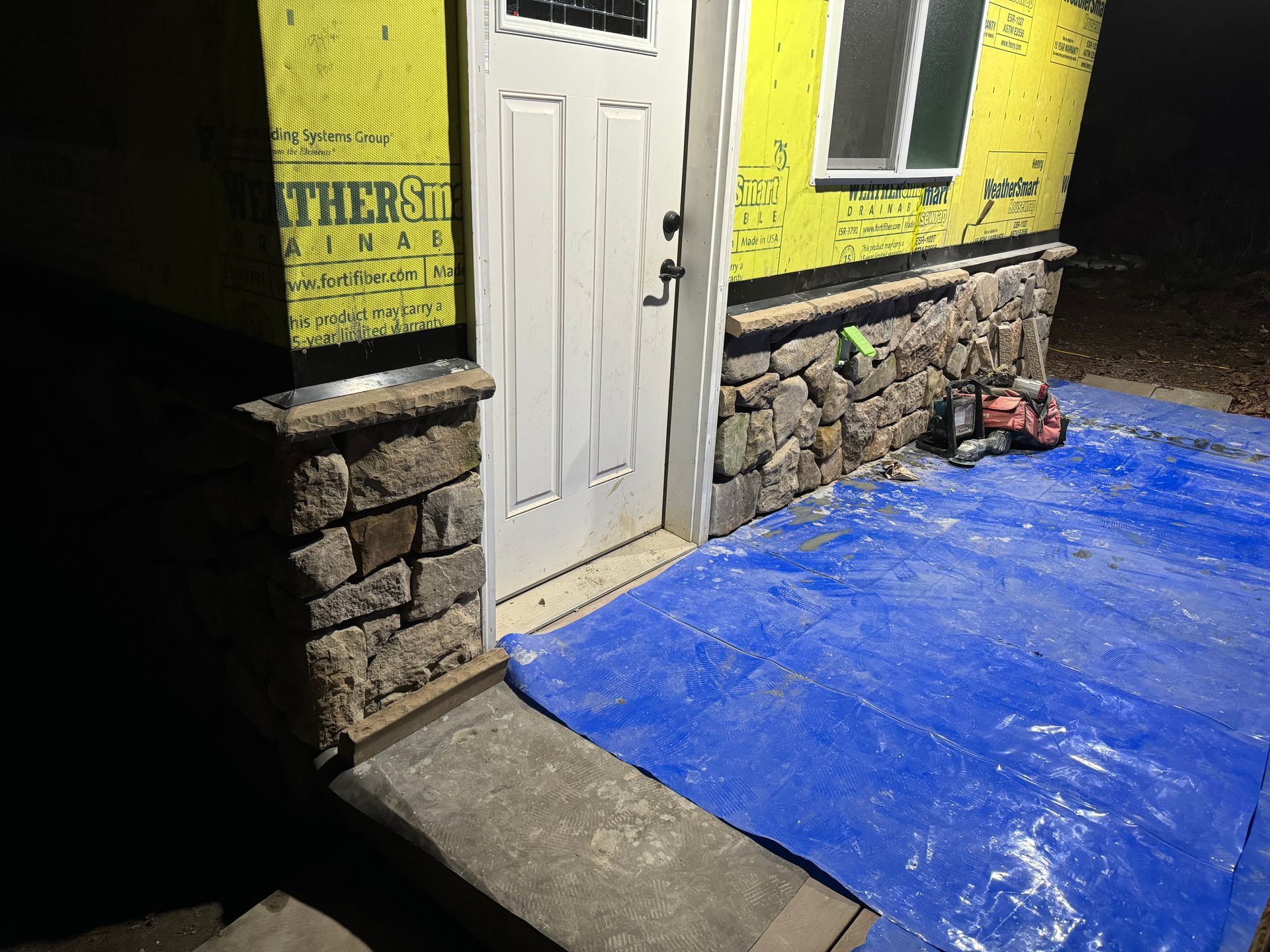 Stone facade construction around a white door. A blue tarp covers the ground.