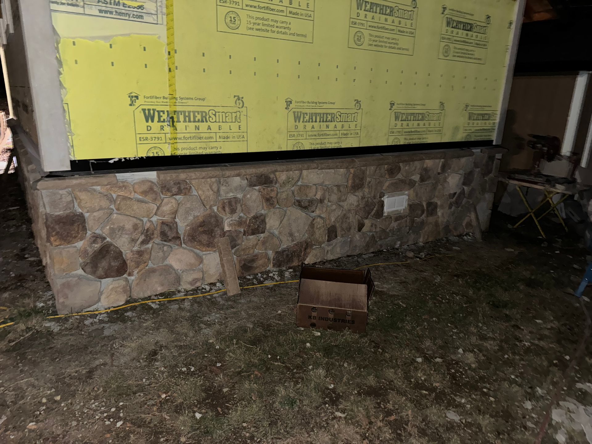 Stone foundation of a building with weathered yellow siding above. Brown cardboard box sits on the ground.