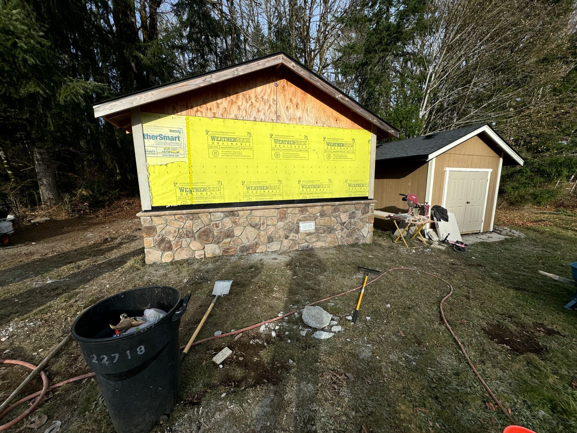 A building under construction with a stone base and yellow insulation, surrounded by tools and a trash can.