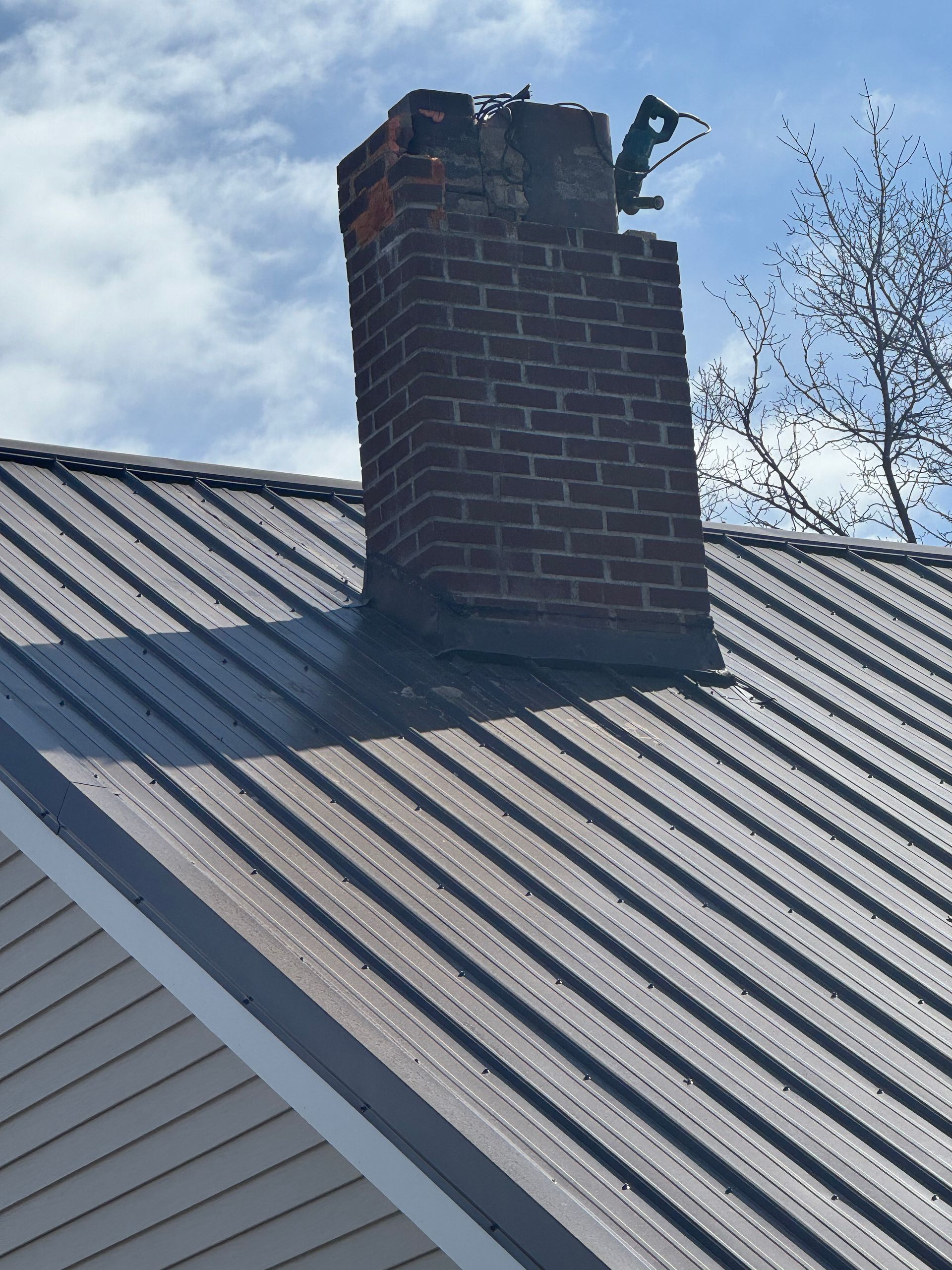 Brick chimney on a metal roof, with damage near the top and a sky background.
