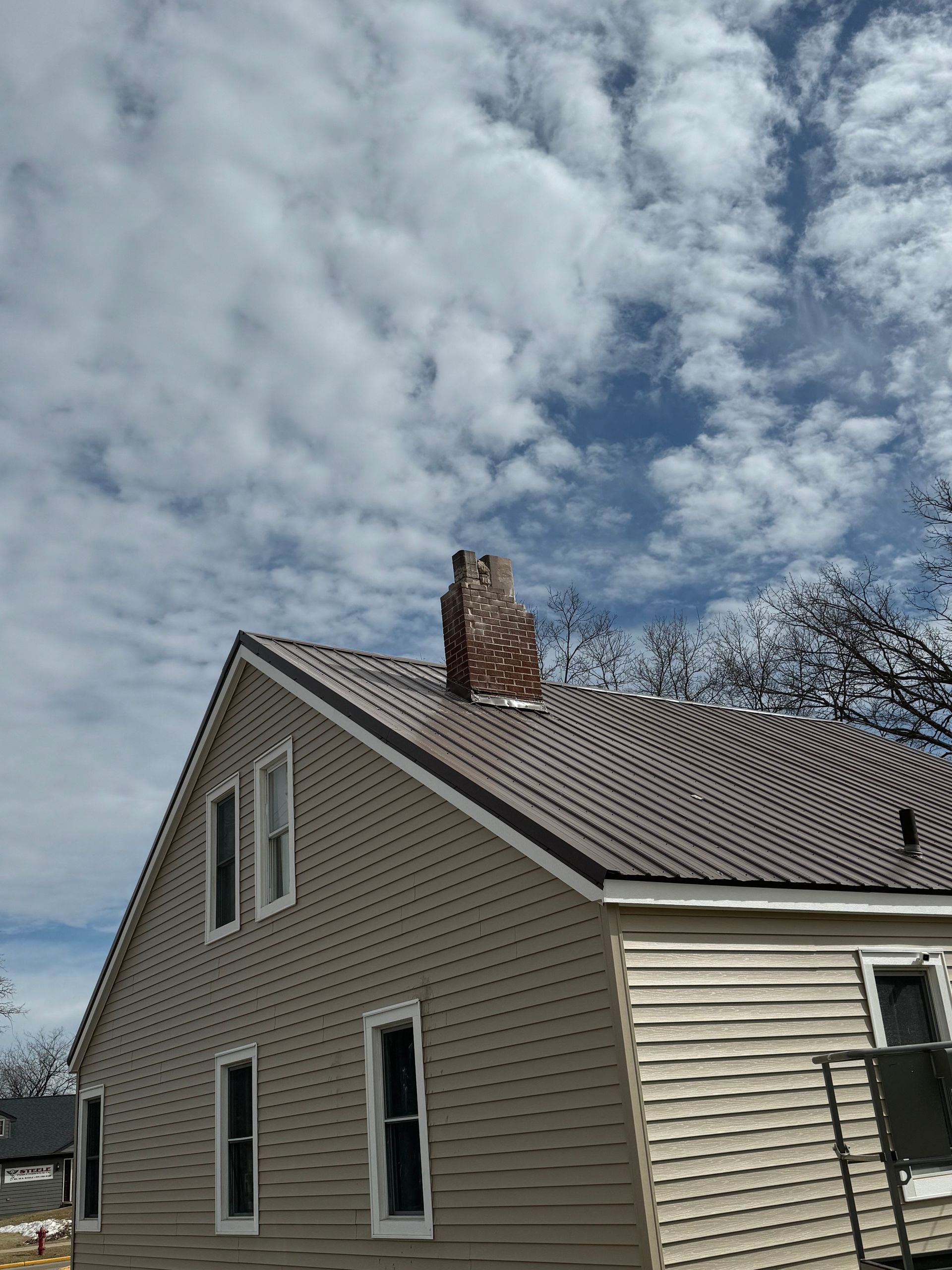 Tan house with a brown roof and chimney against a cloudy blue sky.