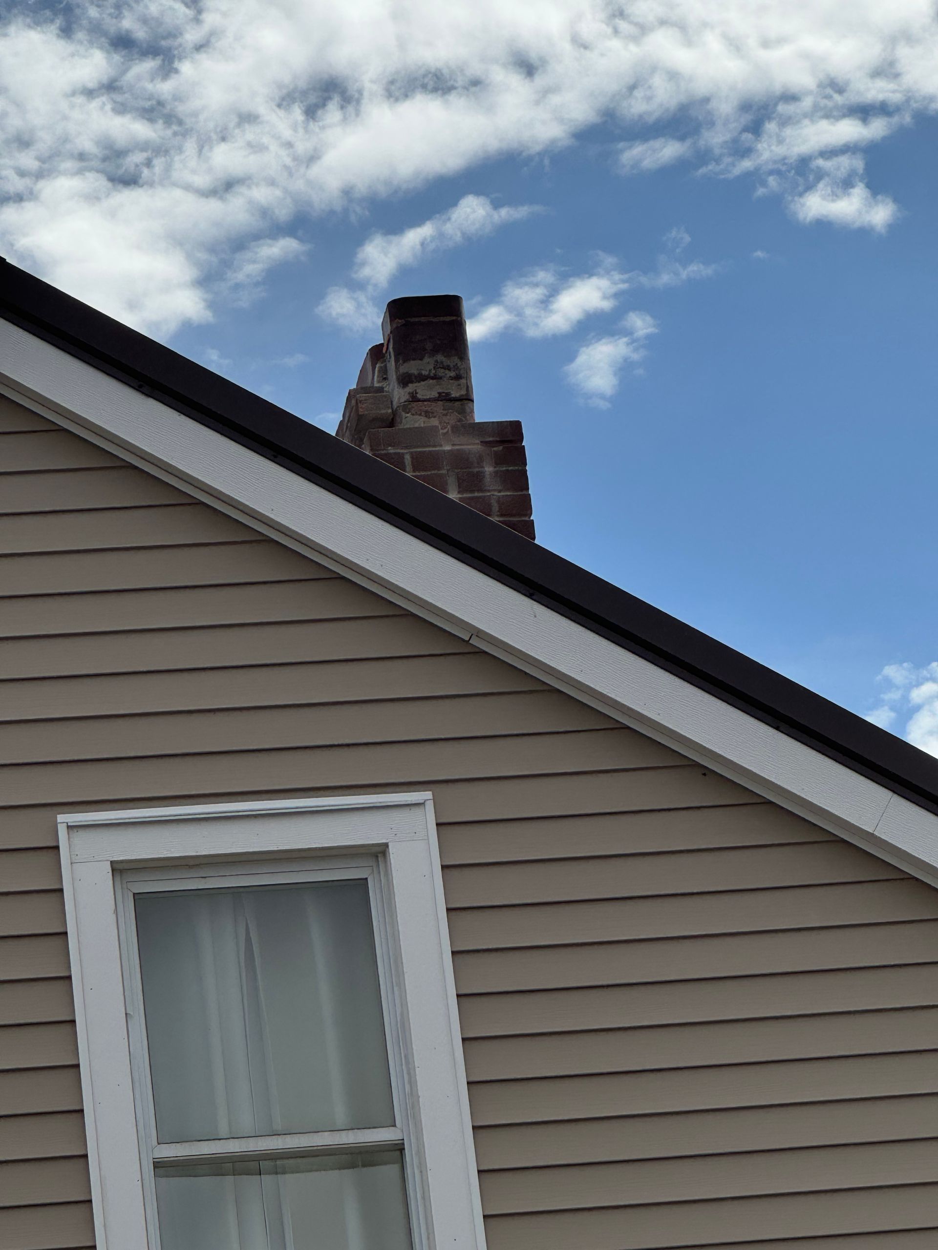 Brick chimney on a beige house with a dark roof against a blue sky with clouds.
