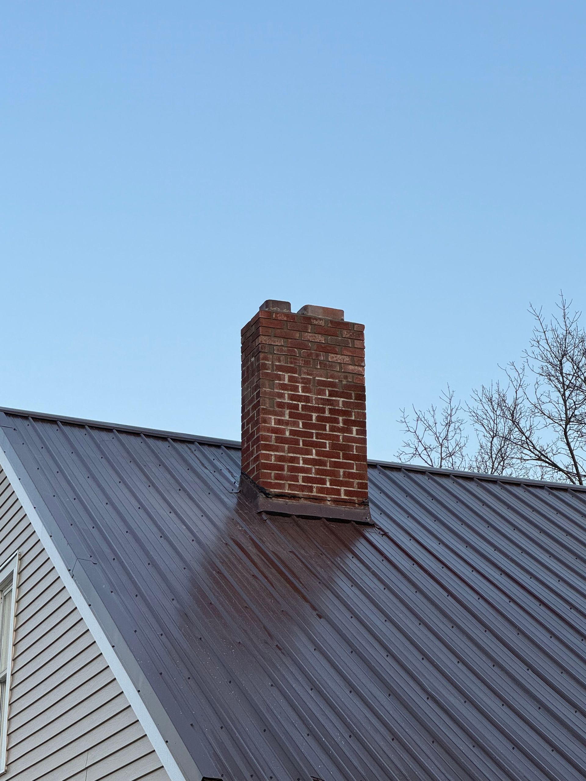 Brick chimney on a dark brown metal roof against a blue sky.