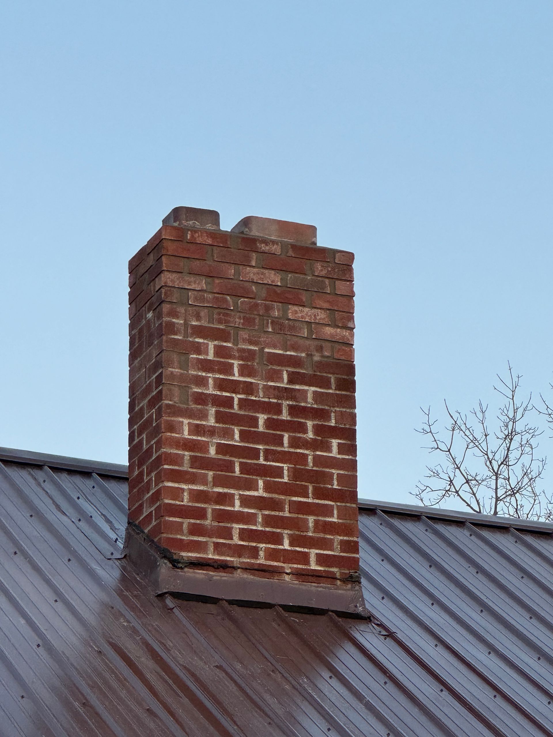 Brick chimney on a dark metal roof against a clear blue sky.