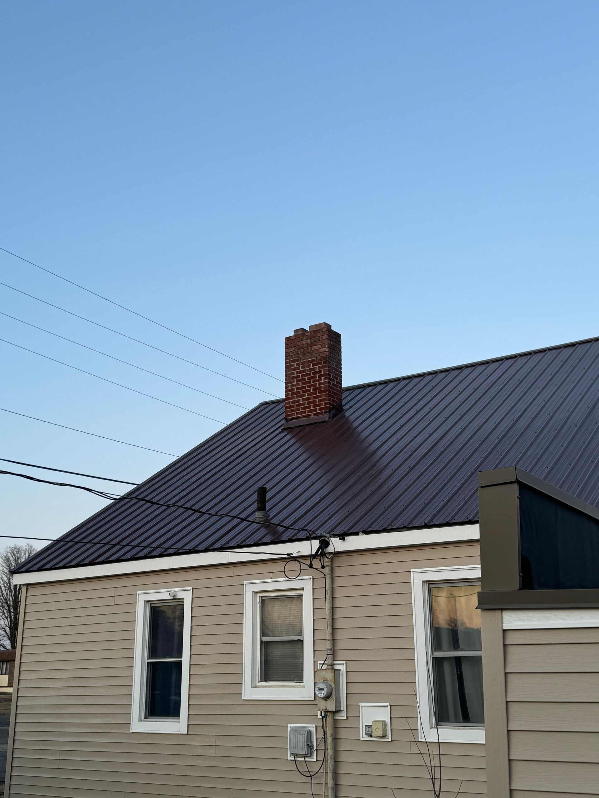 Tan house with dark metal roof, brick chimney, and clear blue sky.