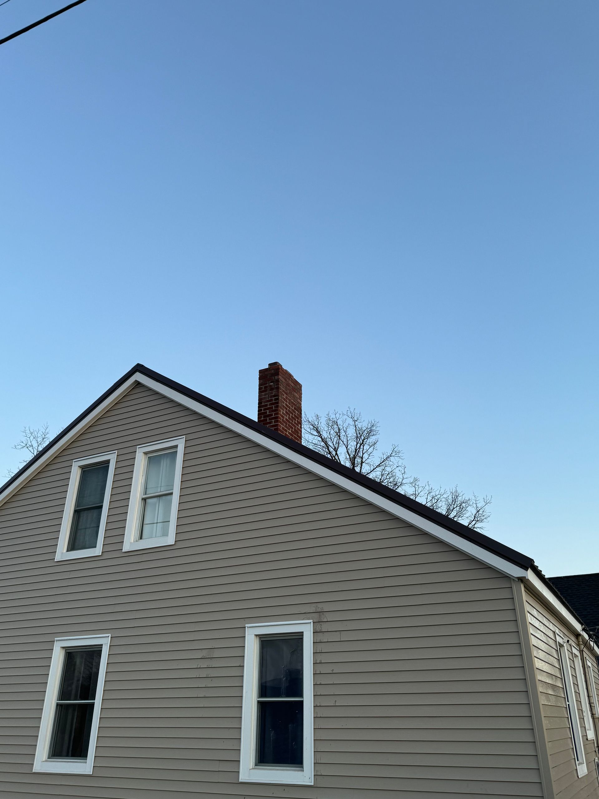 Two-story house with beige siding, black roof, brick chimney, and blue sky.