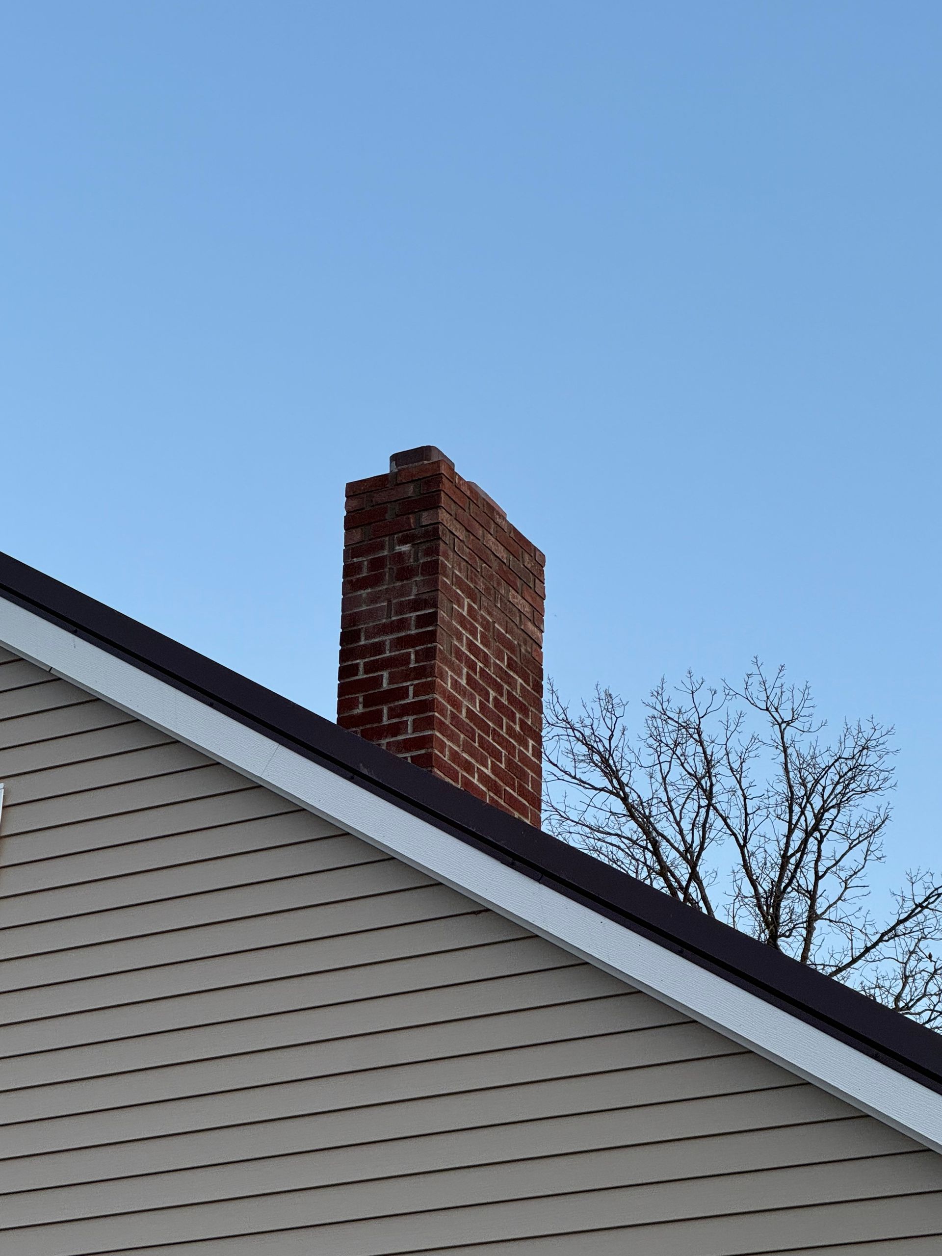 Brick chimney atop a house roof against a clear blue sky.