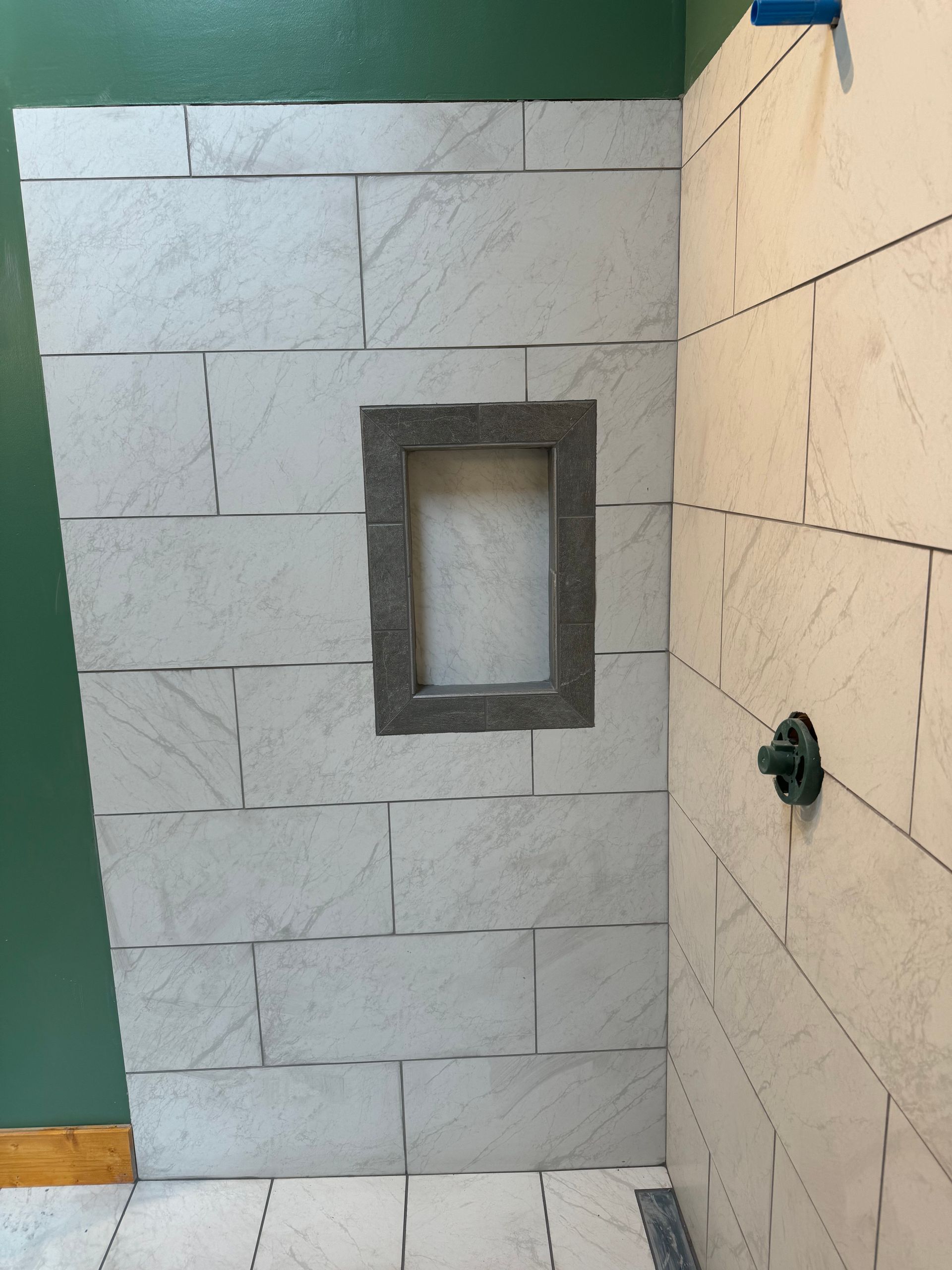 Shower wall with gray tiled niche and shower head. Light gray tiles with a wood-like pattern.
