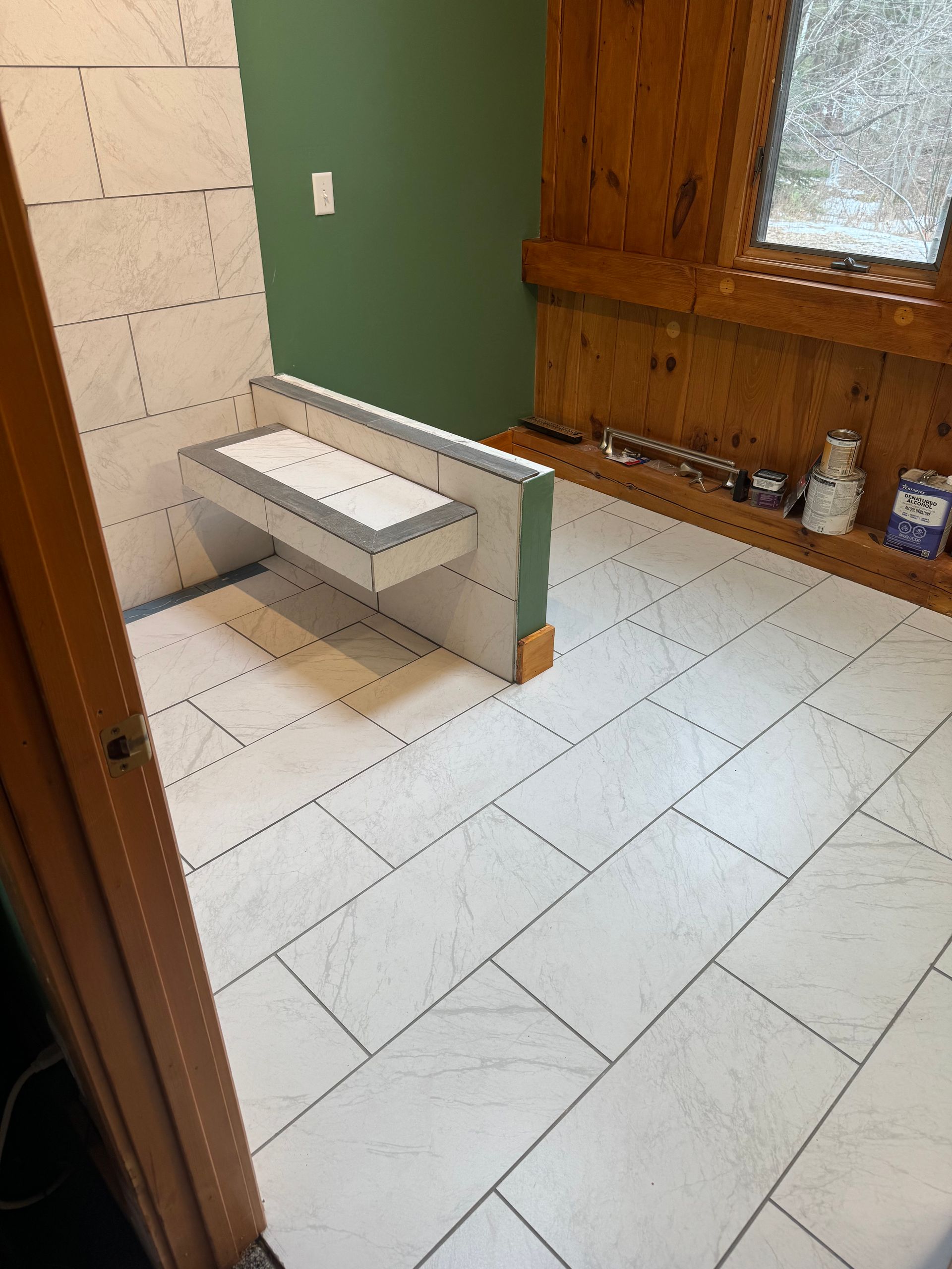 Bathroom with white tiled floor, shower walls, and a built-in bench. Green wall and wood trim.