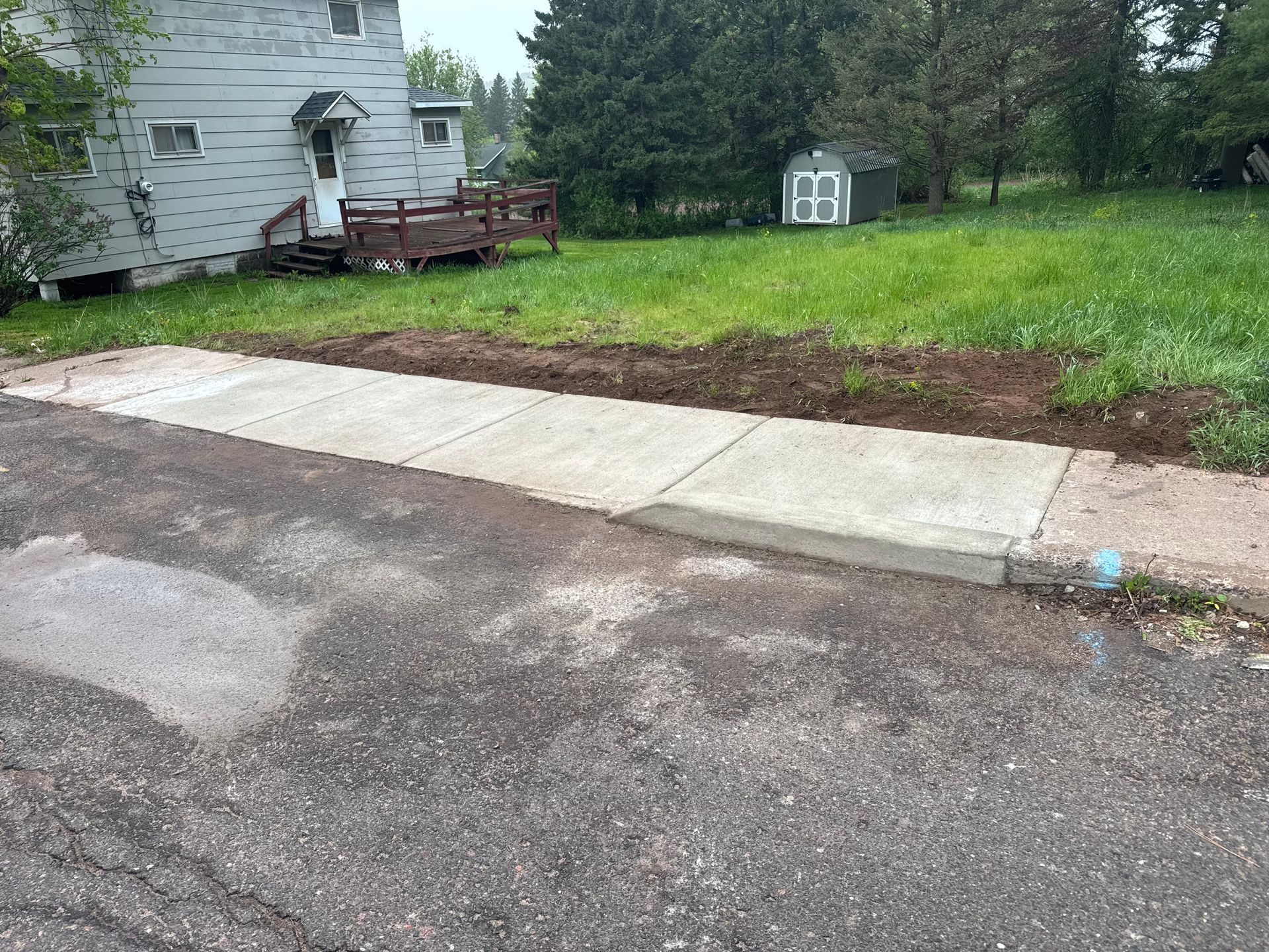 Concrete sidewalk and driveway border a grassy yard with a house and shed in the background.