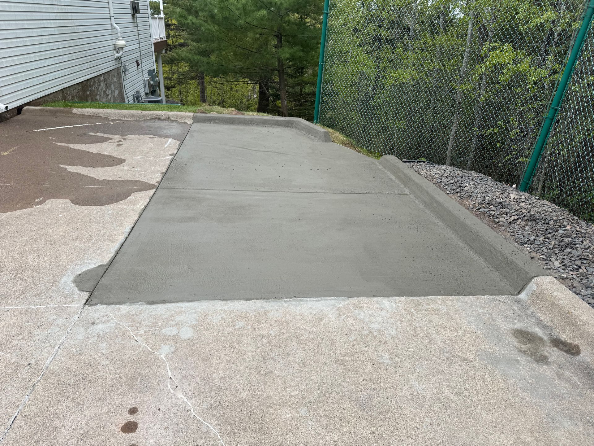 Freshly poured concrete pad adjacent to a building, bordered by existing concrete and a fence.