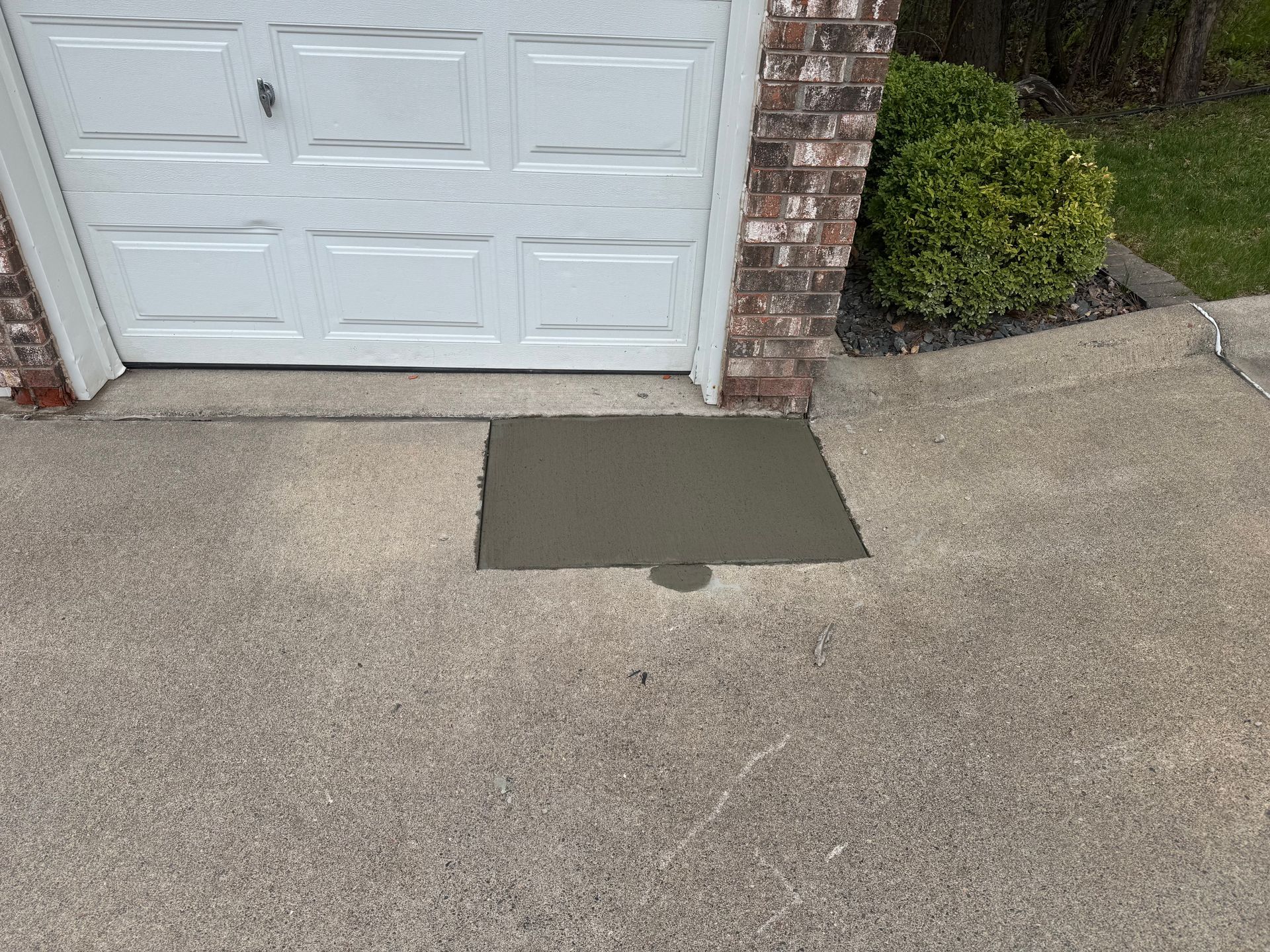 Freshly poured concrete patch on a textured driveway in front of a white garage door.