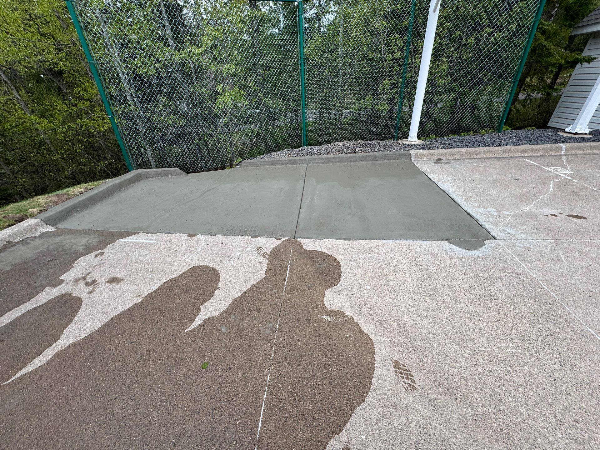 Freshly poured concrete patch on a driveway next to an older concrete surface, near a chain-link fence.