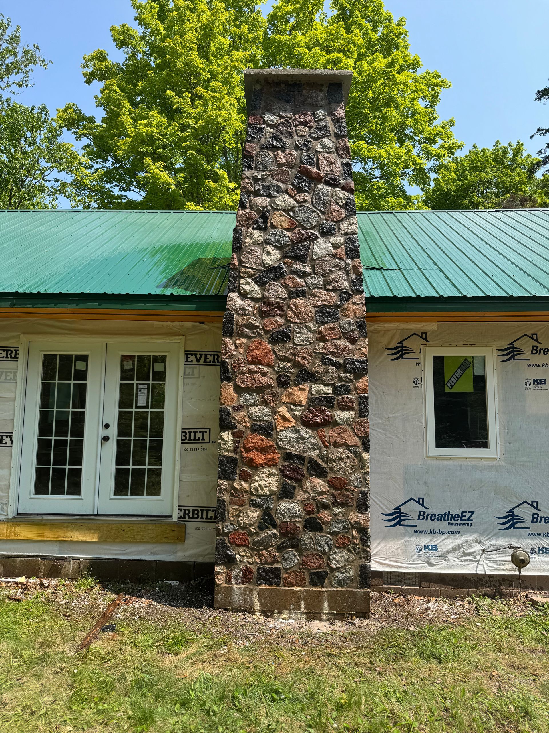 Stone chimney on a building with a green roof, two doors, and a window; construction in progress.