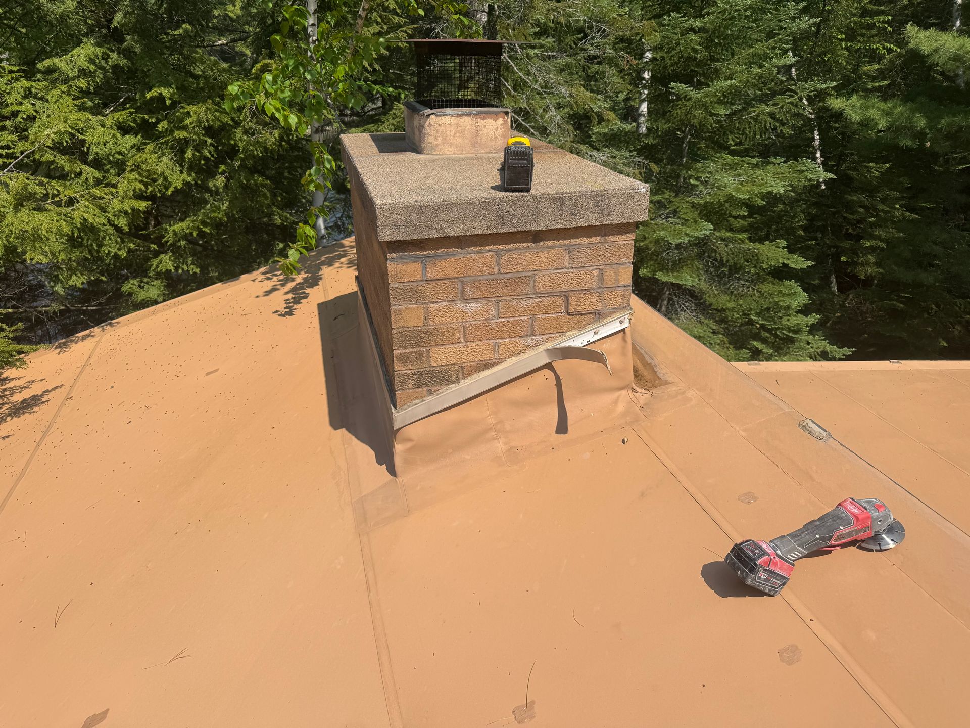 Chimney on a brown metal roof with a grinder on the right, surrounded by trees.