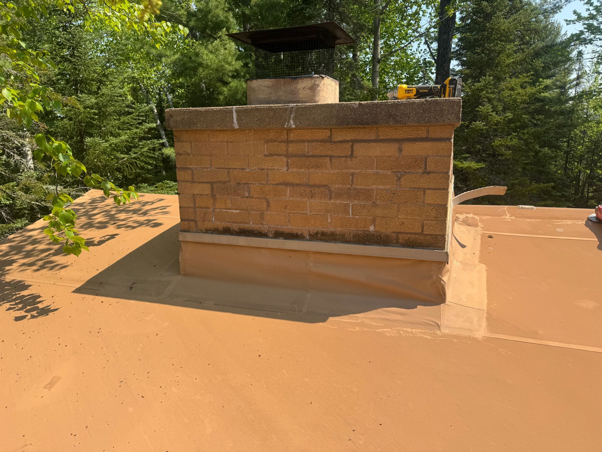 Brick chimney on a brown roof, surrounded by green trees.