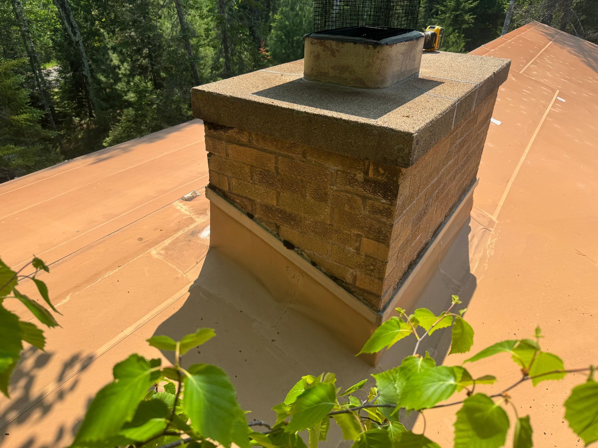 Brick chimney on a brown roof, surrounded by green foliage, in a sunny outdoor setting.