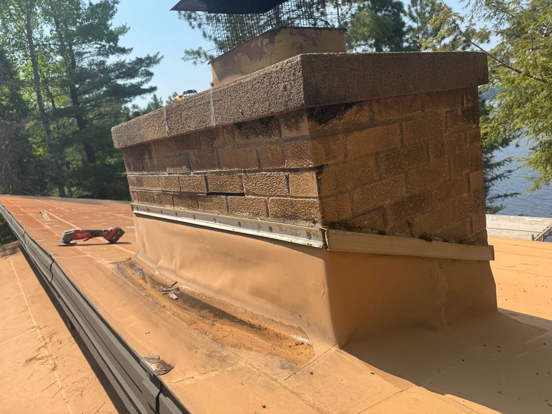 Chimney on a roof. Brick structure with flashing, showing weathering. Sunny, outdoor setting.