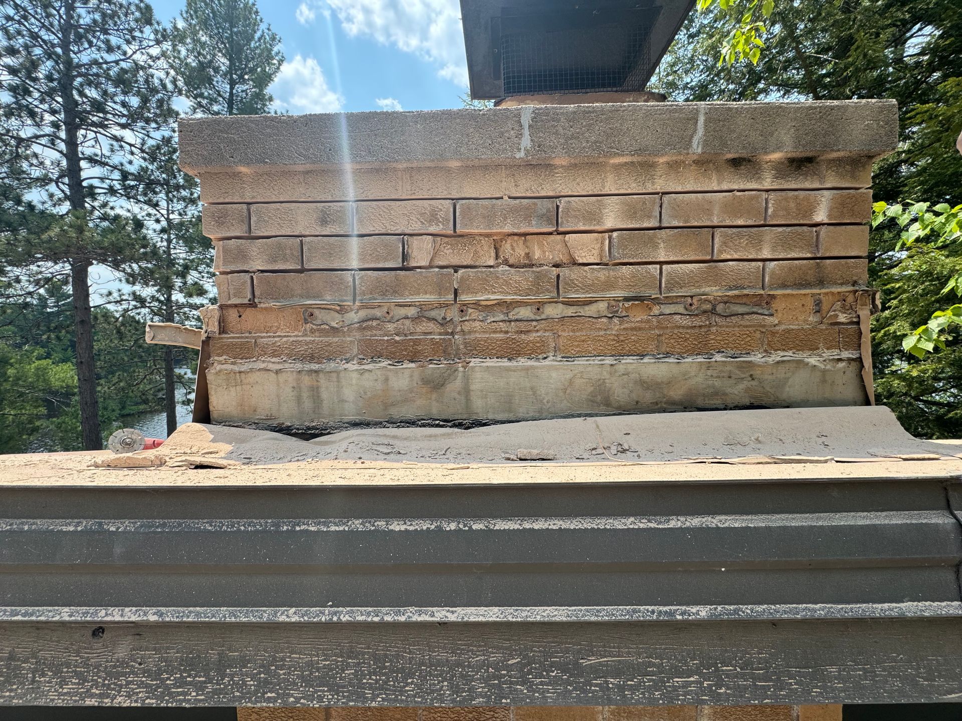 A deteriorating brick chimney on a roof with a damaged mortar, set against a backdrop of trees and a bright sky.