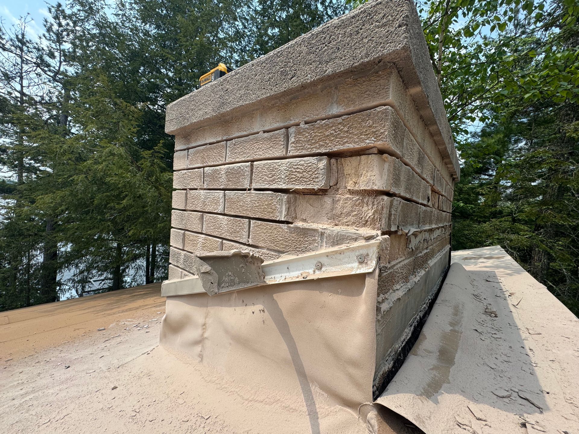 Brick chimney with visible cracks, built on a roof. Beige bricks, concrete cap, and waterproof membrane visible.