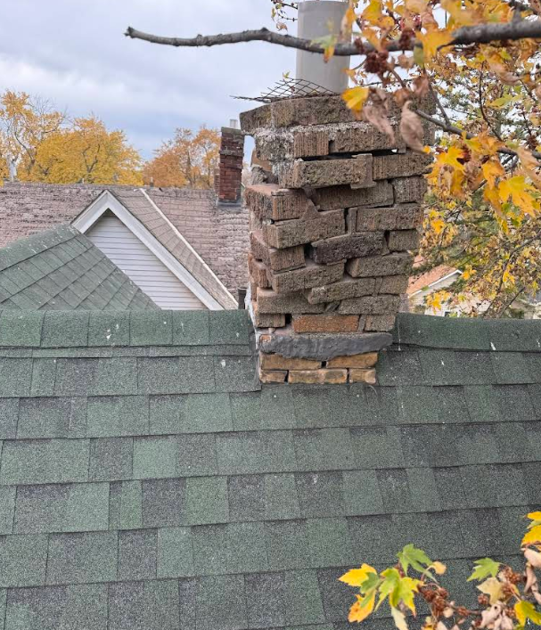 Damaged brick chimney on a green shingled roof, with autumn leaves in the background.