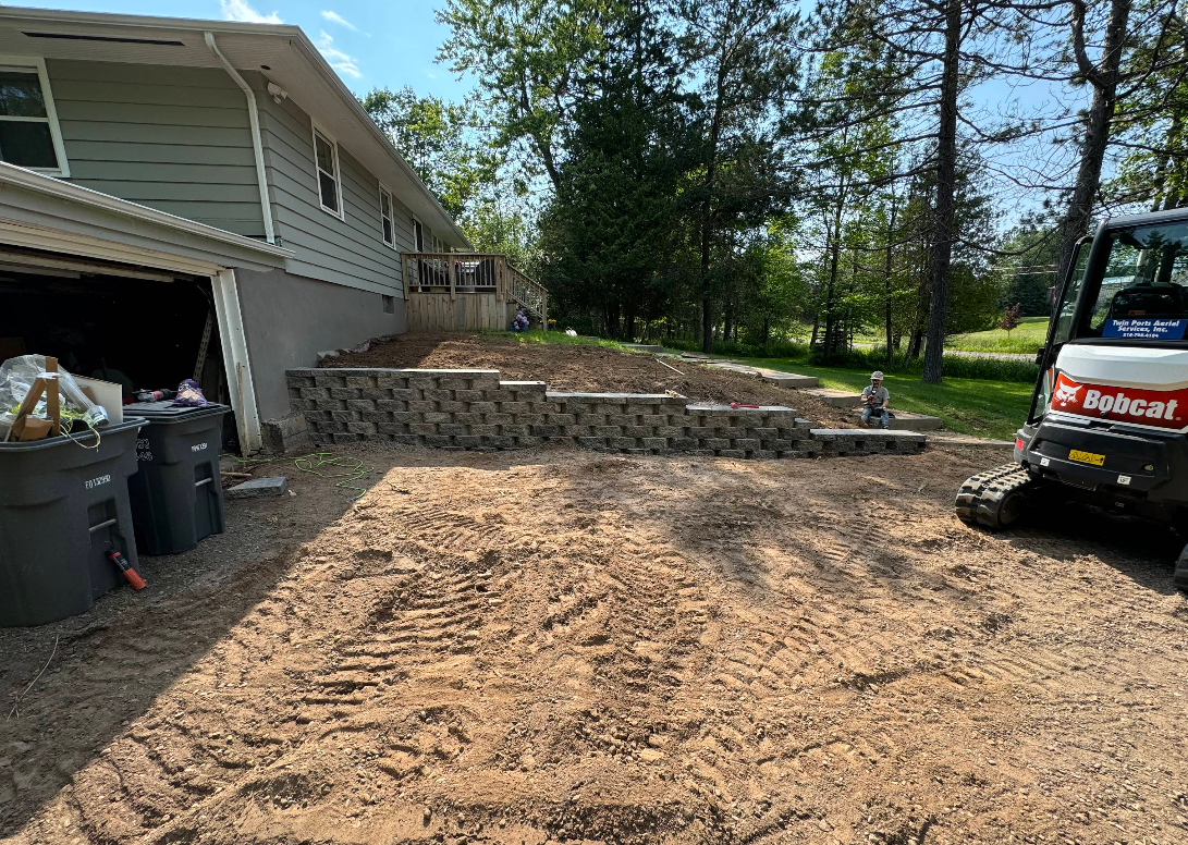 A retaining wall and steps being built near a house. A small excavator sits to the right, in a dirt yard.