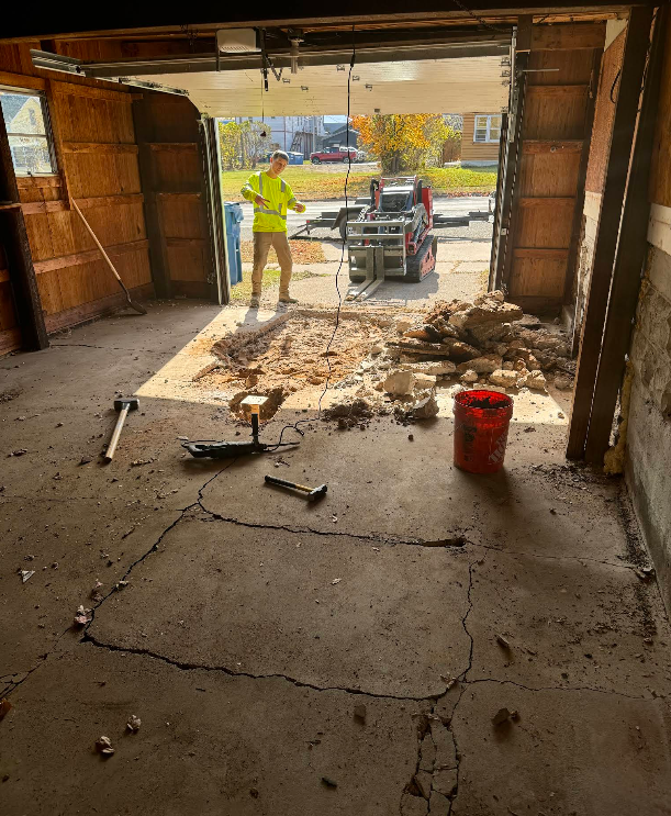 Man operating machinery in garage with debris. Door open to outside with vehicle and trees.