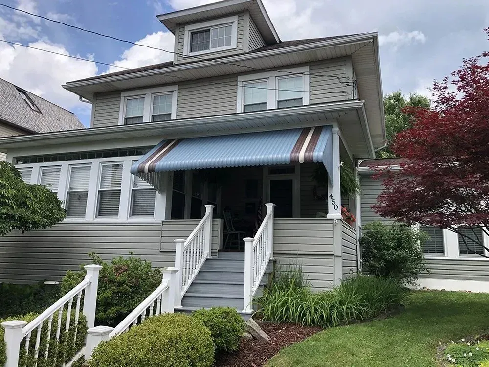 Two-story gray house with white trim, porch, and blue awning. White railing leads to front door.