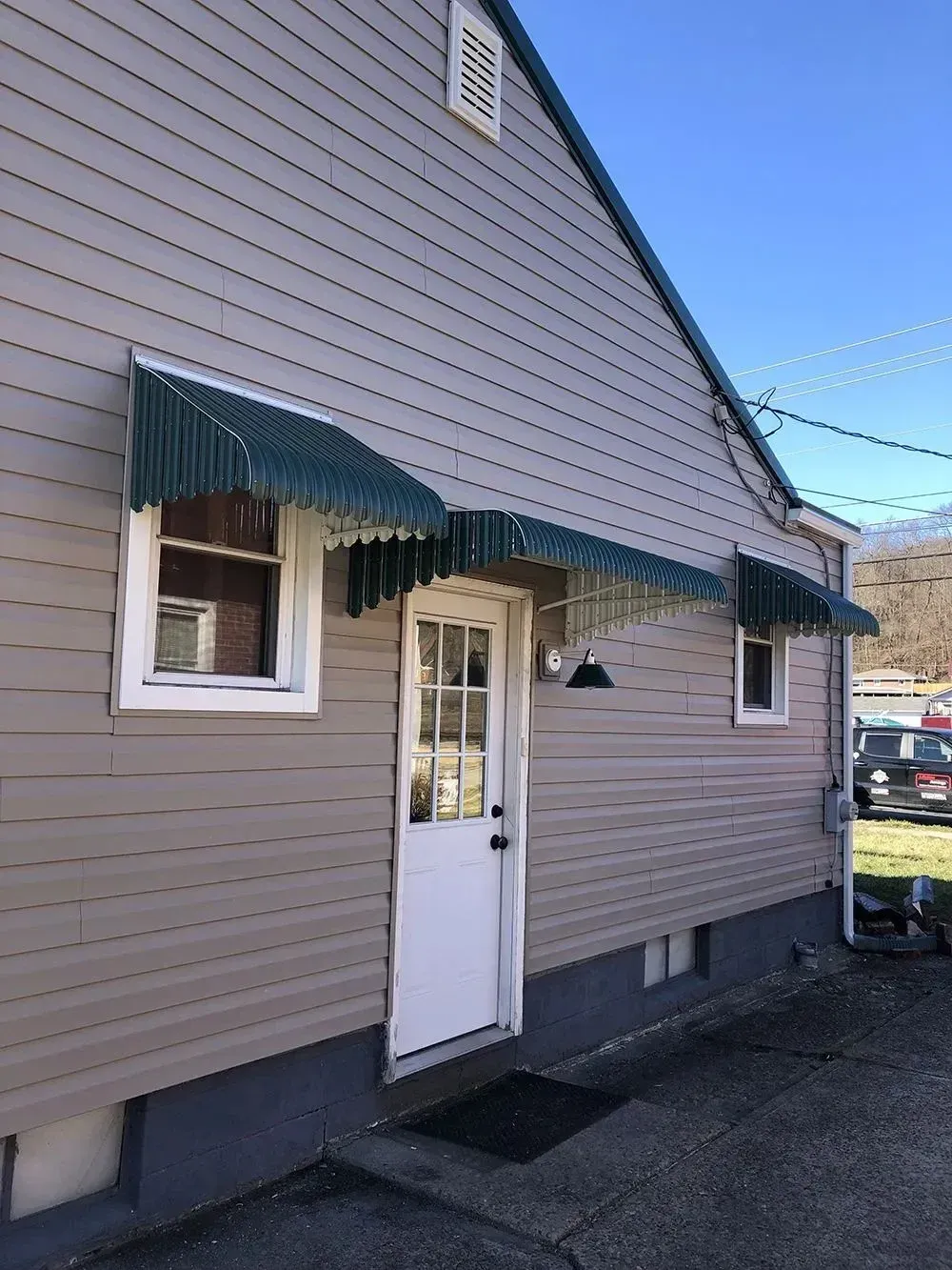 Side view of a building with a white door, windows, and green awnings. Tan siding and a clear blue sky.
