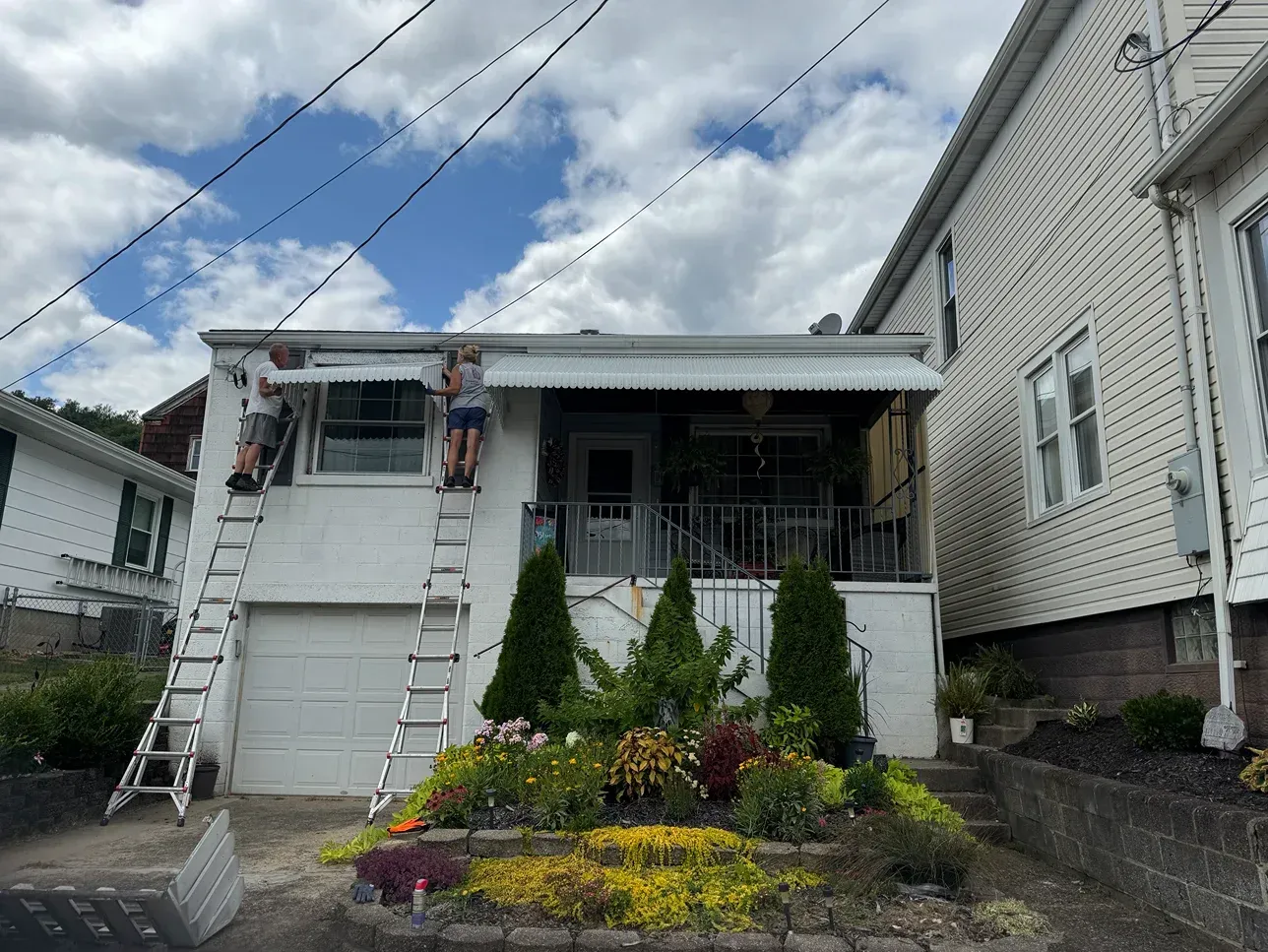 Two people on ladders painting the white trim of a small house with a colorful garden and a cloudy sky.