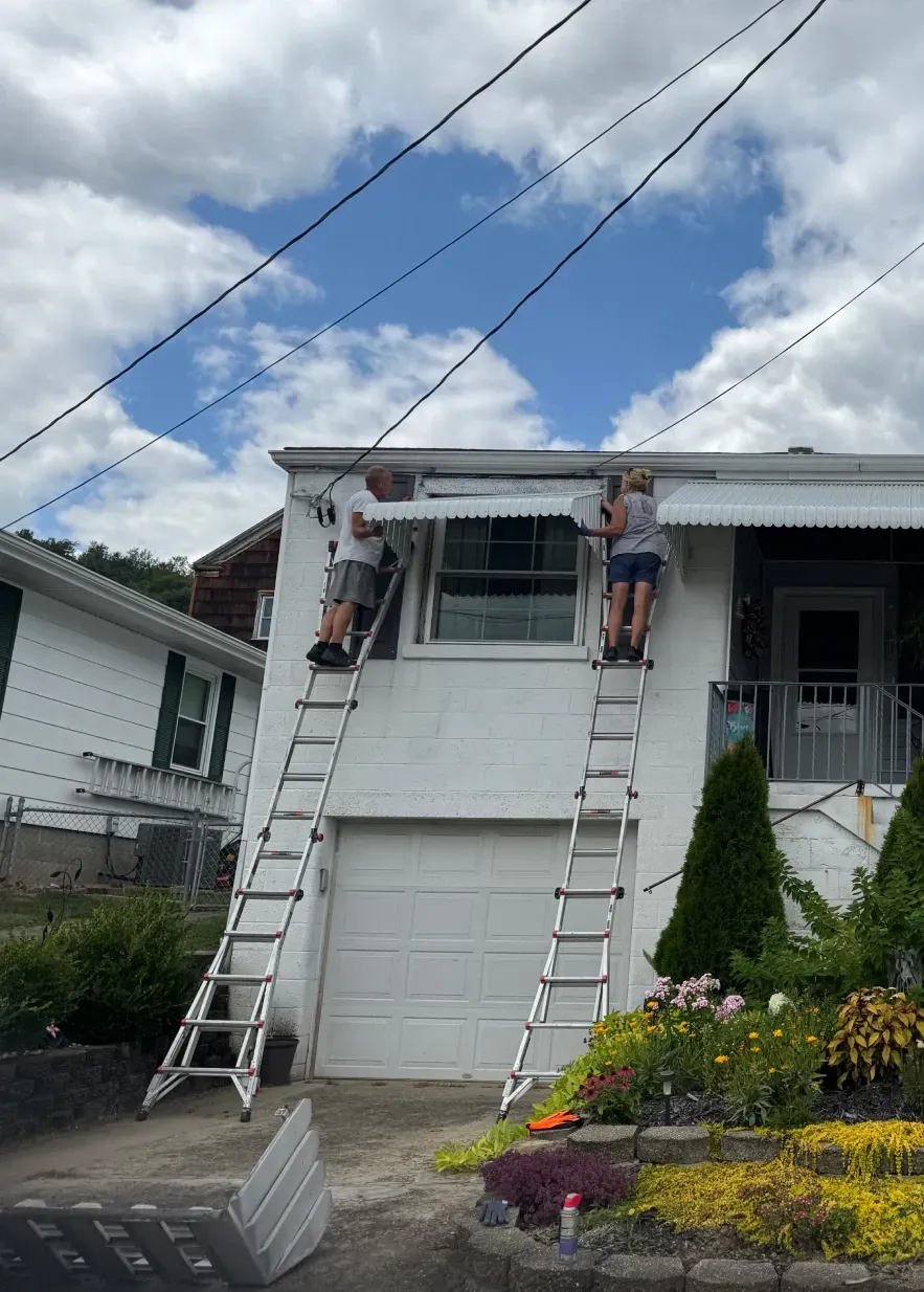 Two people on ladders working on a white building's awning. One person is on the left, one on the right. Blue sky background.