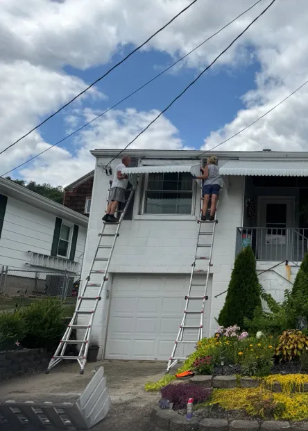 Two people on ladders working on a white building's awning. One person is on the left, one on the right. Blue sky background.