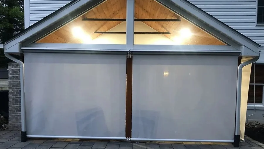Outdoor patio with two large, light-colored roll-down shades under a wooden ceiling; overhead lights illuminate the space.