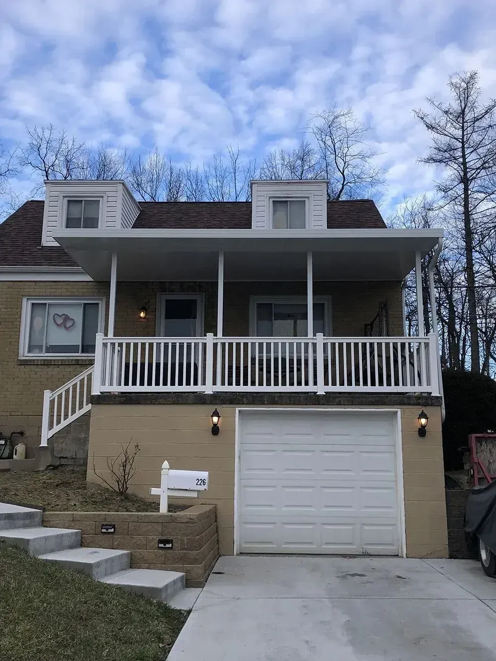 Tan brick house with a white porch, garage, and steps leading to the front door, under a cloudy sky.