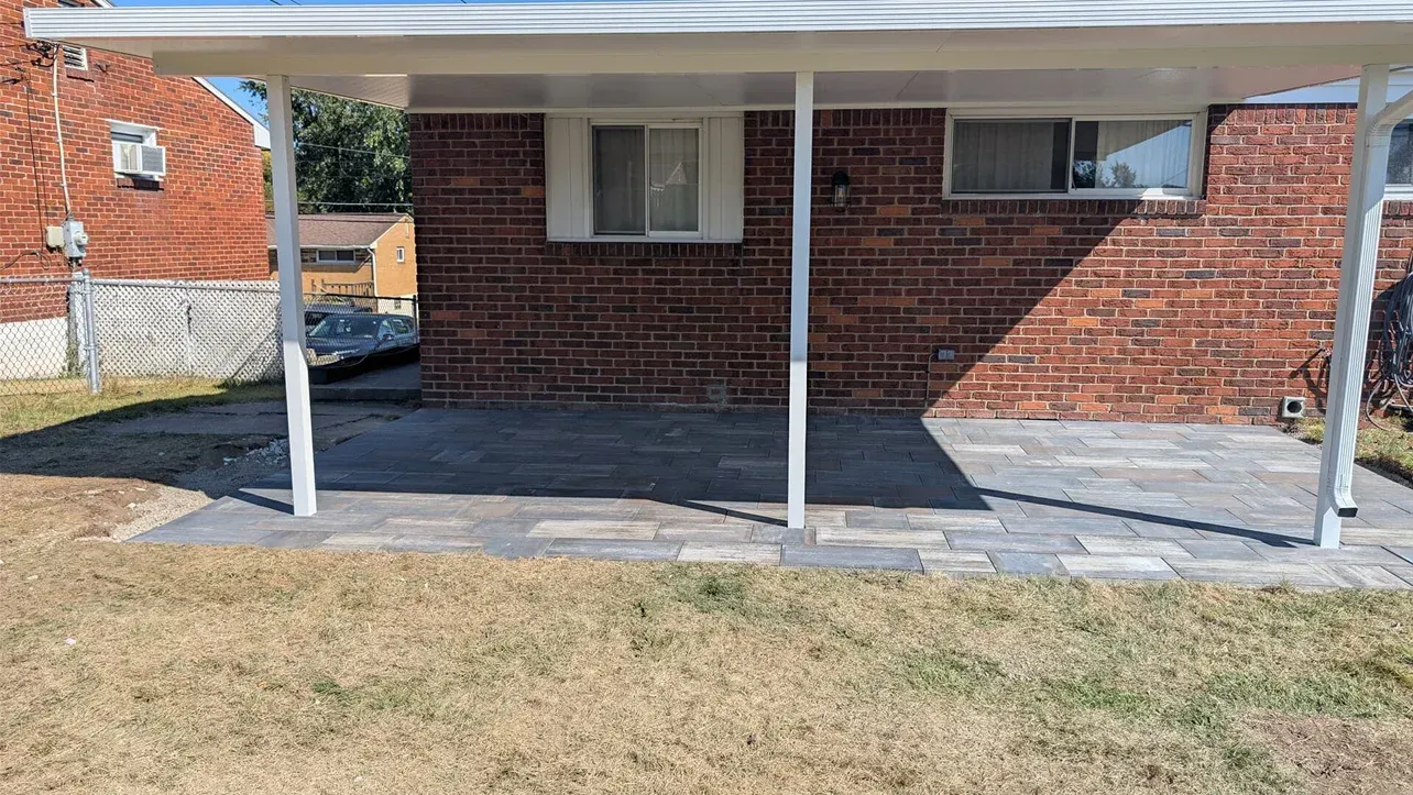 White carport over a brick patio with two windows, supported by white posts, with a grassy lawn in the foreground.