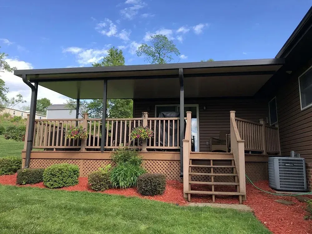 Covered deck with stairs, surrounded by plants and red mulch, attached to a brown house with air conditioning unit.