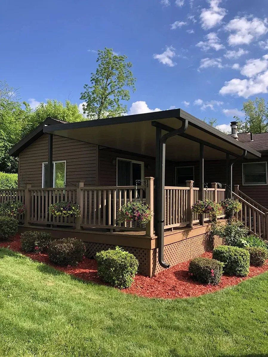 A brown house with a covered deck, surrounded by greenery and red mulch under a blue sky.