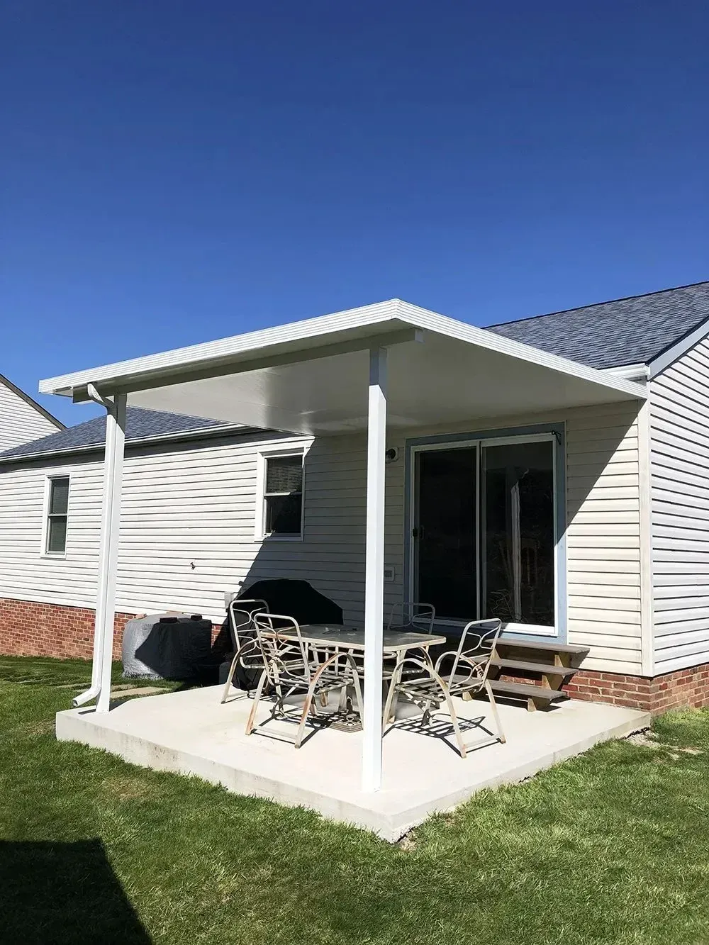 Patio with a white covered pergola, table, and chairs. Next to a house with a sliding glass door.